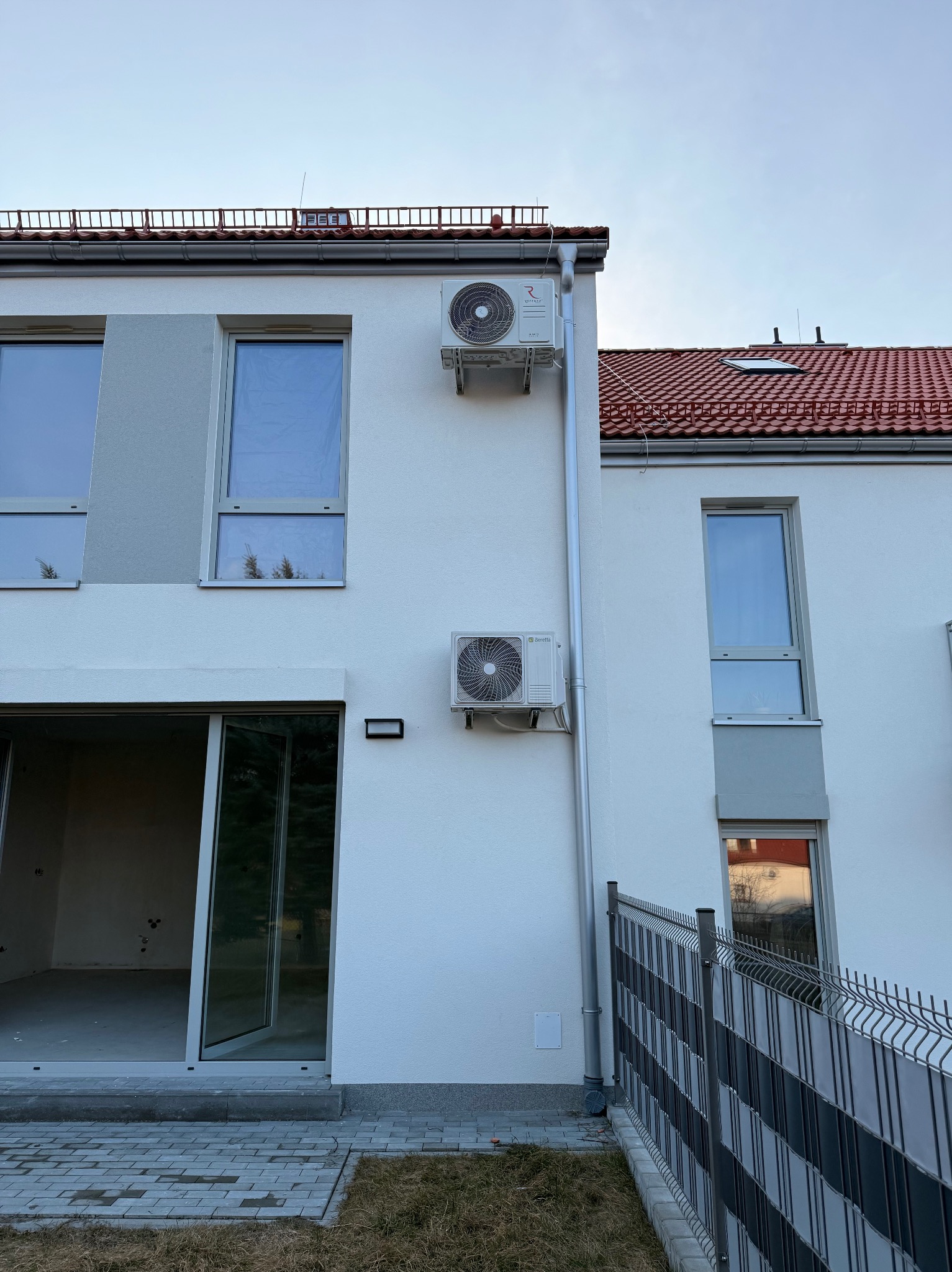 Dwa klimatyzatory na elewacji nowoczesnego, dwukondygnacyjnego domu z czerwonym dachem. Widoczne okna i przesuwne drzwi balkonowe. Częściowo widoczne ogrodzenie.