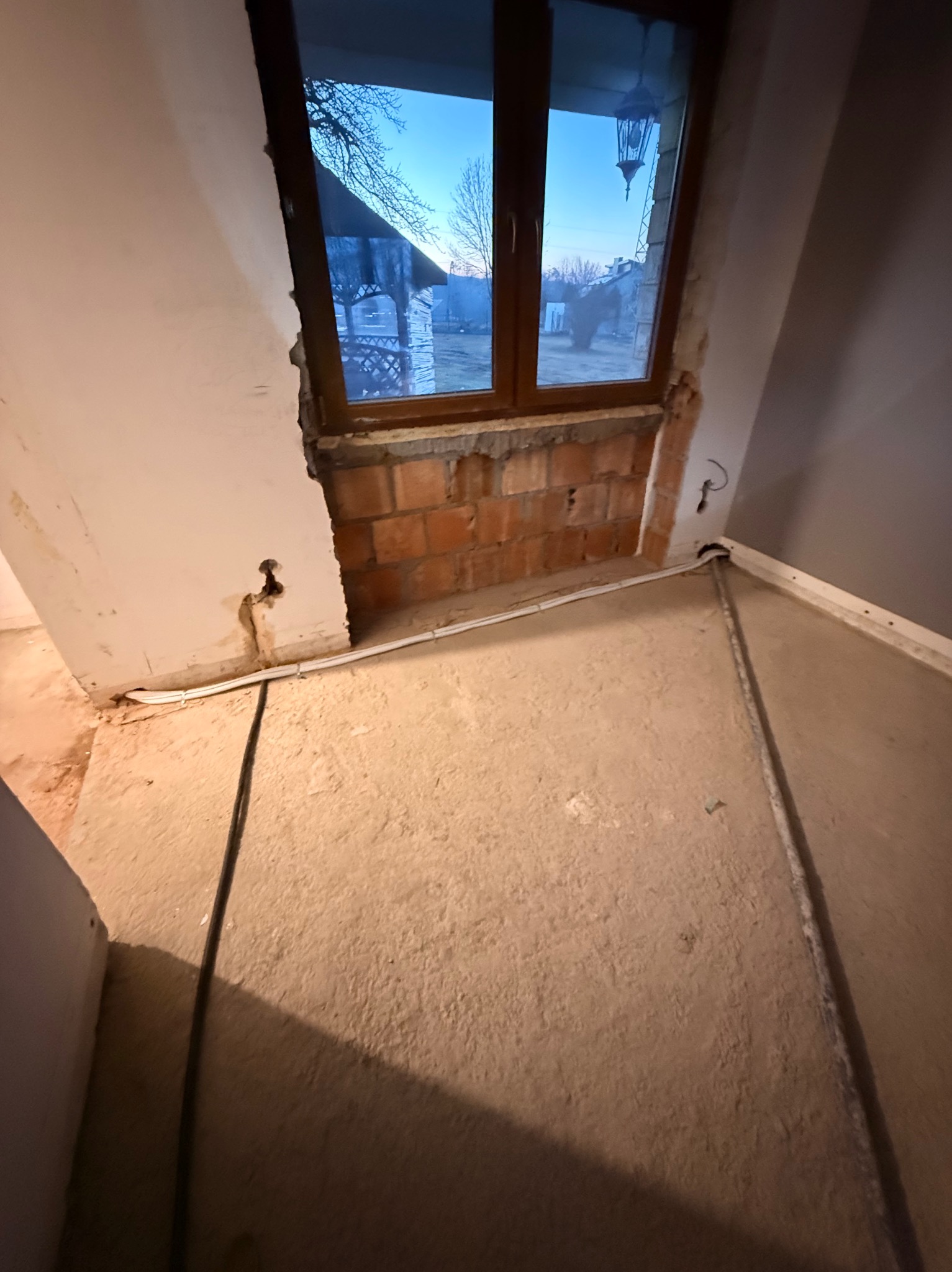 Wnętrze w trakcie remontu: odsłonięta cegła wokół okna, widoczne przewody elektryczne wzdłuż ściany i podłogi. Surowy stan przed wykończeniem.