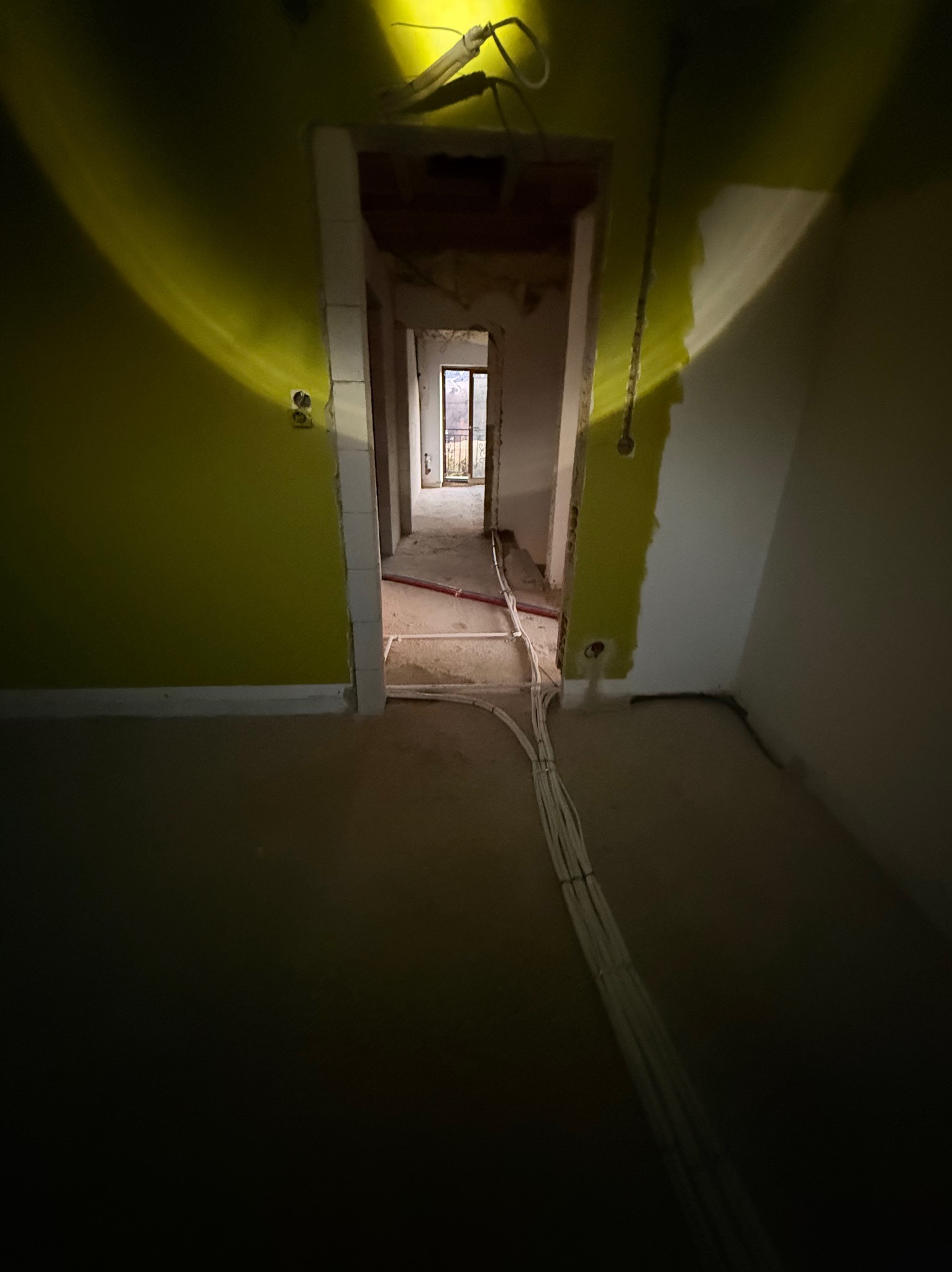Wnętrze w trakcie remontu z widocznymi przewodami elektrycznymi ułożonymi na podłodze, oświetlone punktowo. Ściany w surowym stanie, widoczne otwory na gniazdka.
