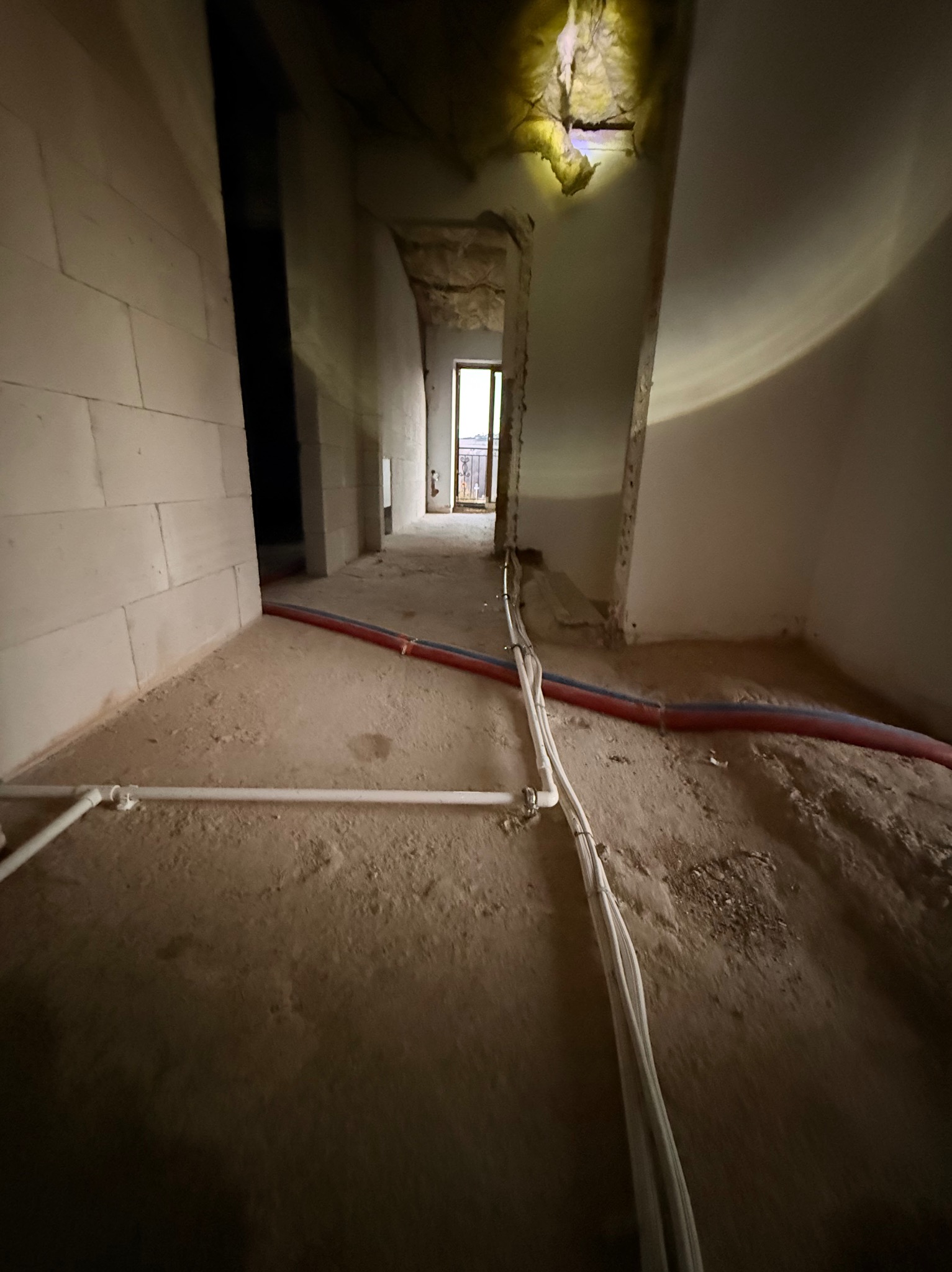 Wnętrze w stanie surowym z widocznymi rurami instalacji wodnej (czerwona i niebieska) oraz przewodami elektrycznymi ułożonymi na podłodze. Ściany z białego bloczka.