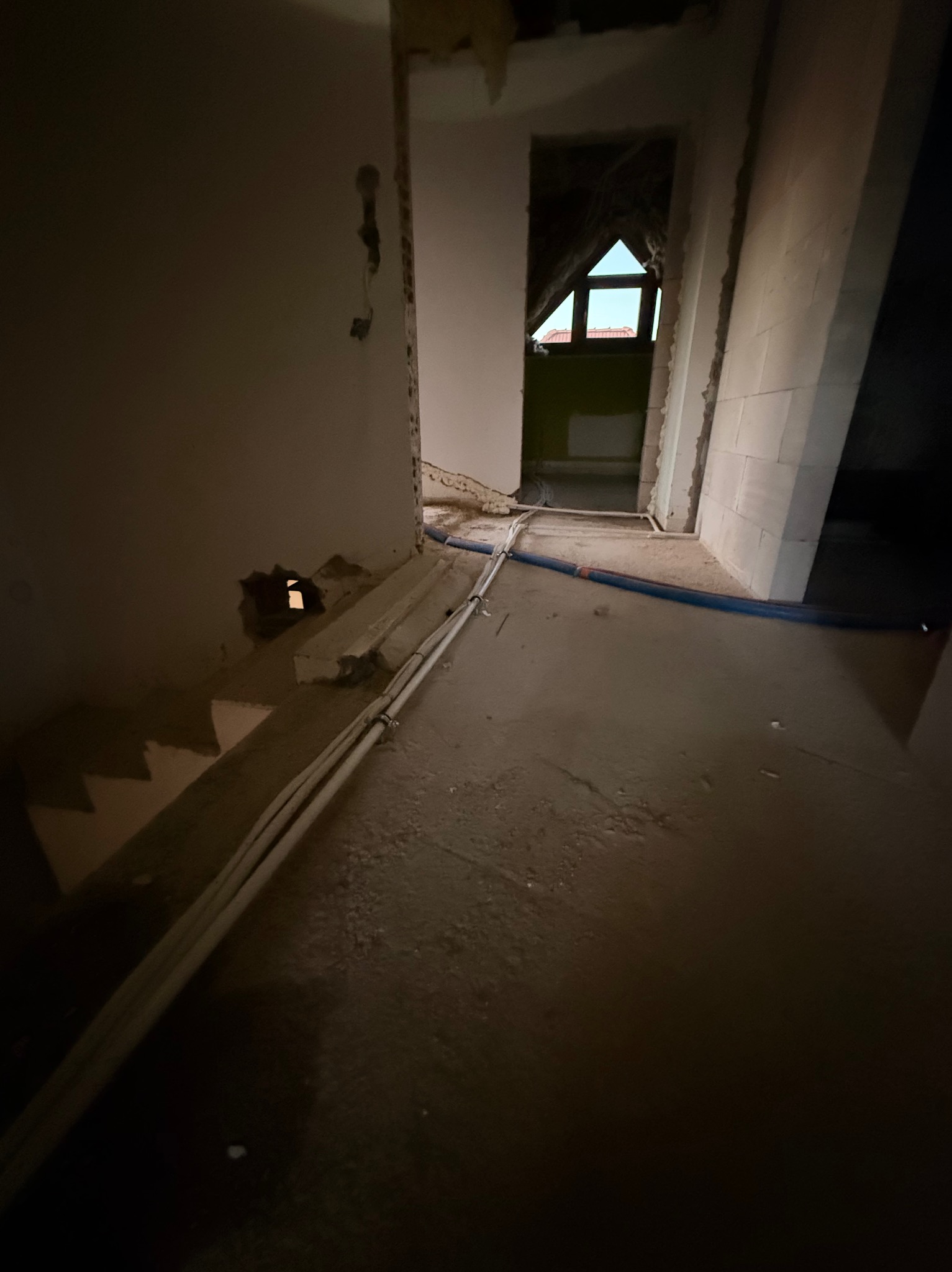 Wnętrze w trakcie remontu: widoczne rury instalacyjne prowadzone wzdłuż ściany, surowy stan wykończenia, okno na poddaszu w tle.