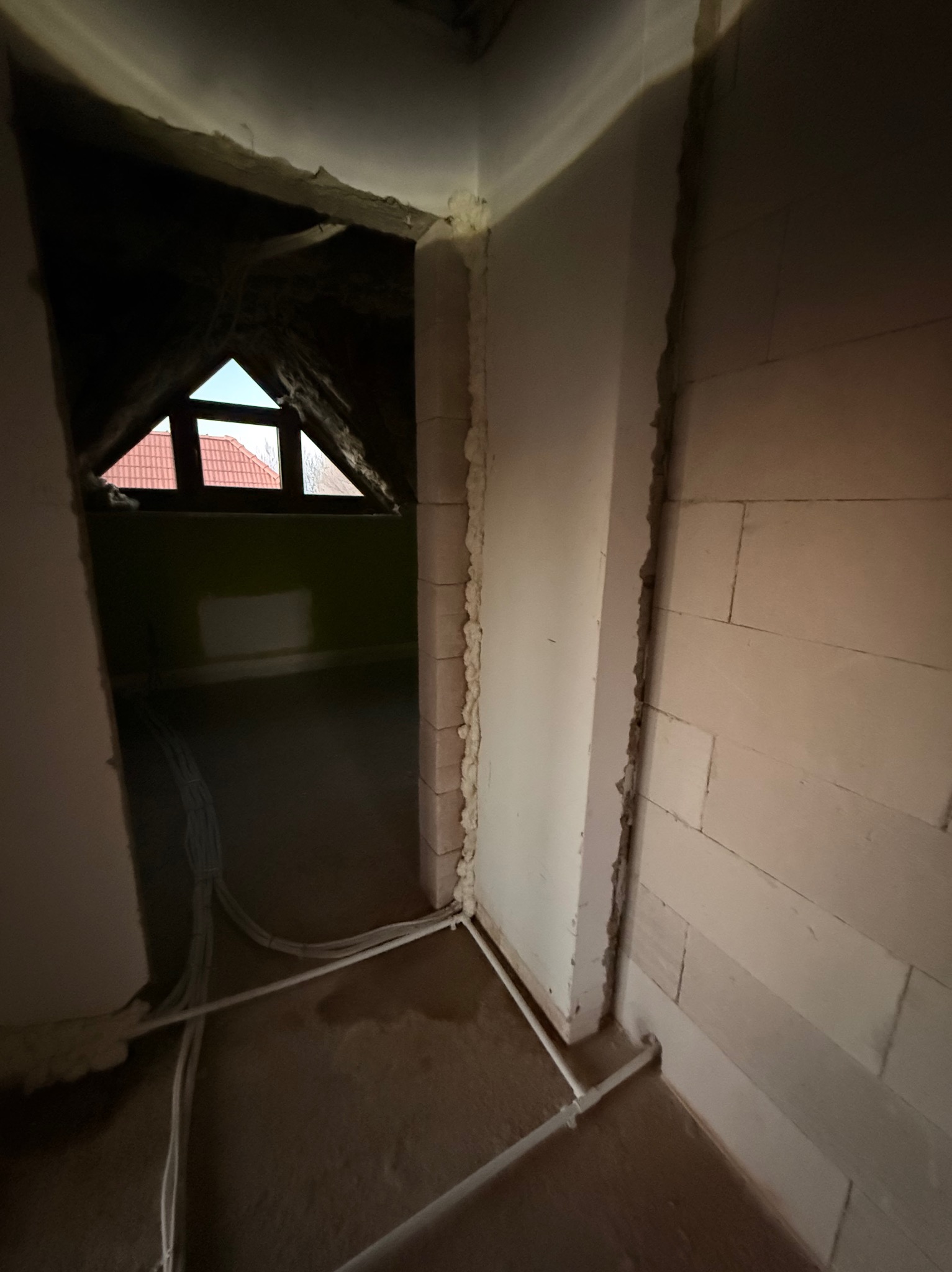 Surowe wnętrze poddasza z oknem, widoczne przewody i rury instalacyjne na podłodze, ściany z bloków. Etap prac instalacyjnych przed wykończeniem.