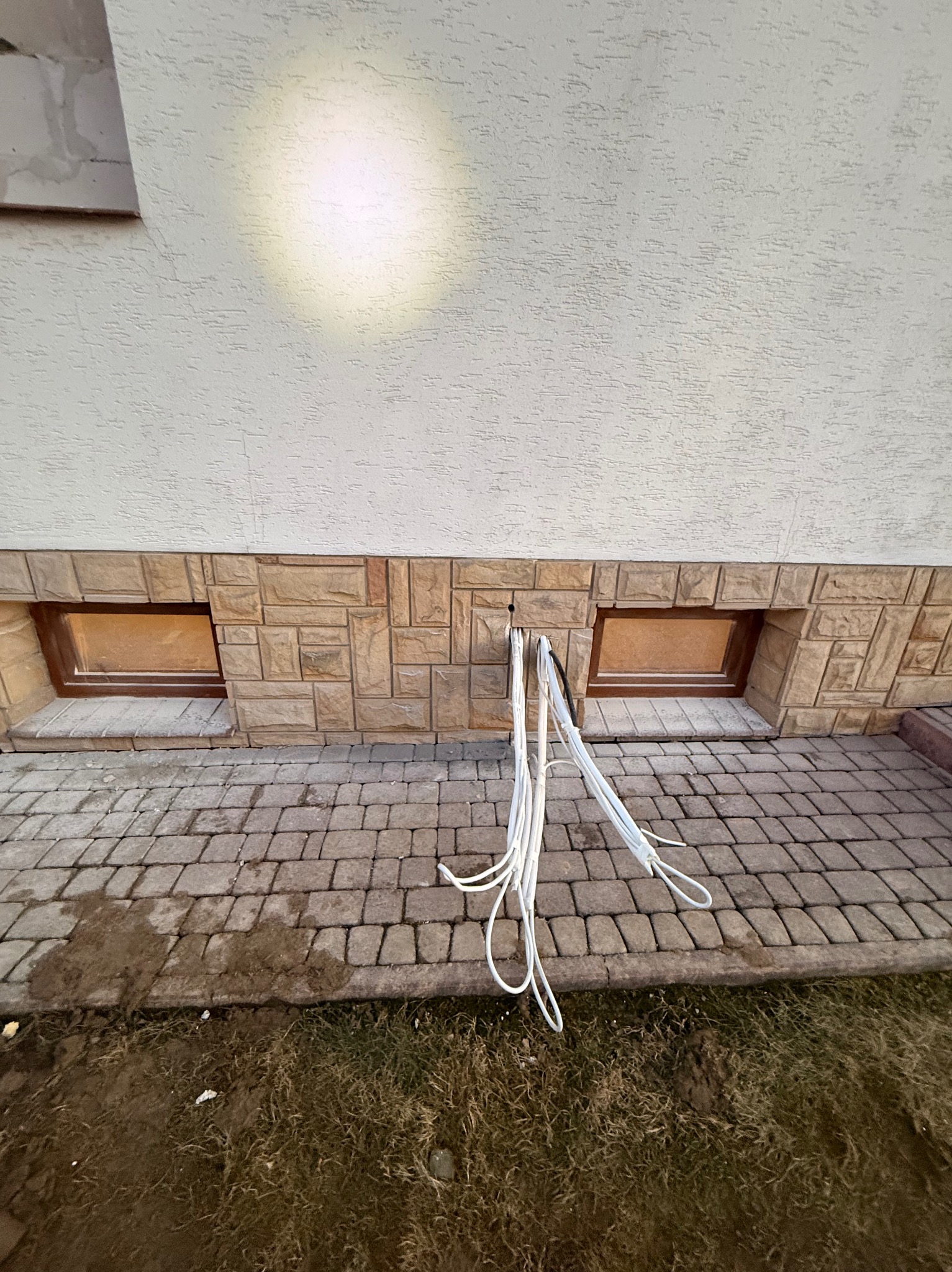 Białe kable wystające ze ściany budynku z oknami piwnicznymi, na tle kostki brukowej i trawy. Instalacja elektryczna w trakcie prac.