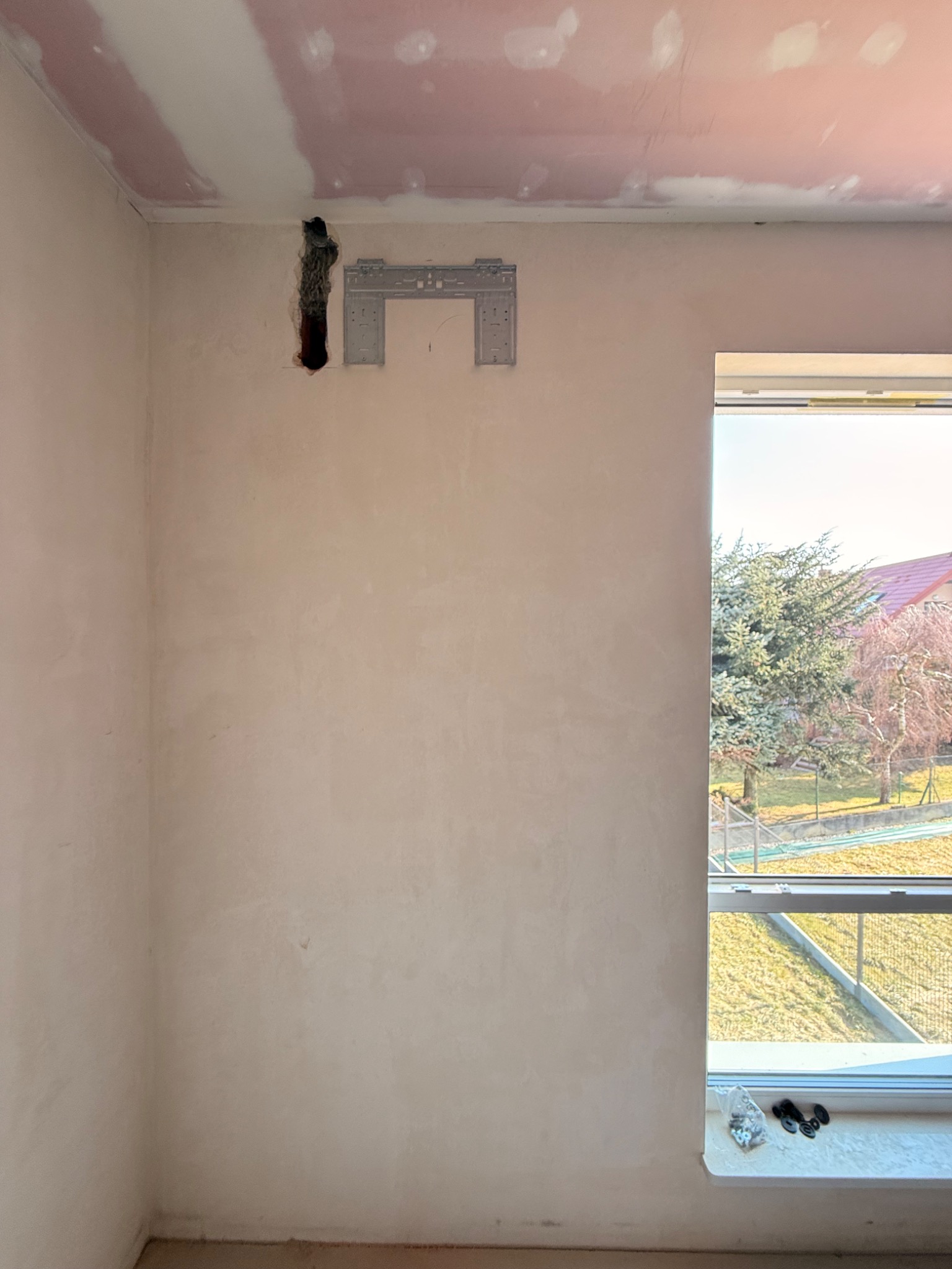 Wnętrze w trakcie montażu klimatyzacji: wspornik na ścianie, otwór na przewody, okno z widokiem na zewnątrz i akcesoria montażowe na parapecie.