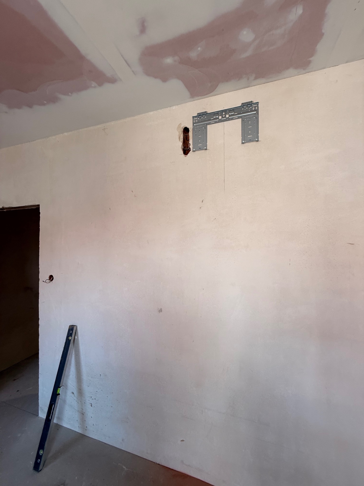 Przygotowanie do montażu klimatyzacji: wspornik na ścianie, rura odpływowa, poziomica przy ścianie. Ściana w trakcie wykończenia, sufit z widocznymi śladami szpachlowania.