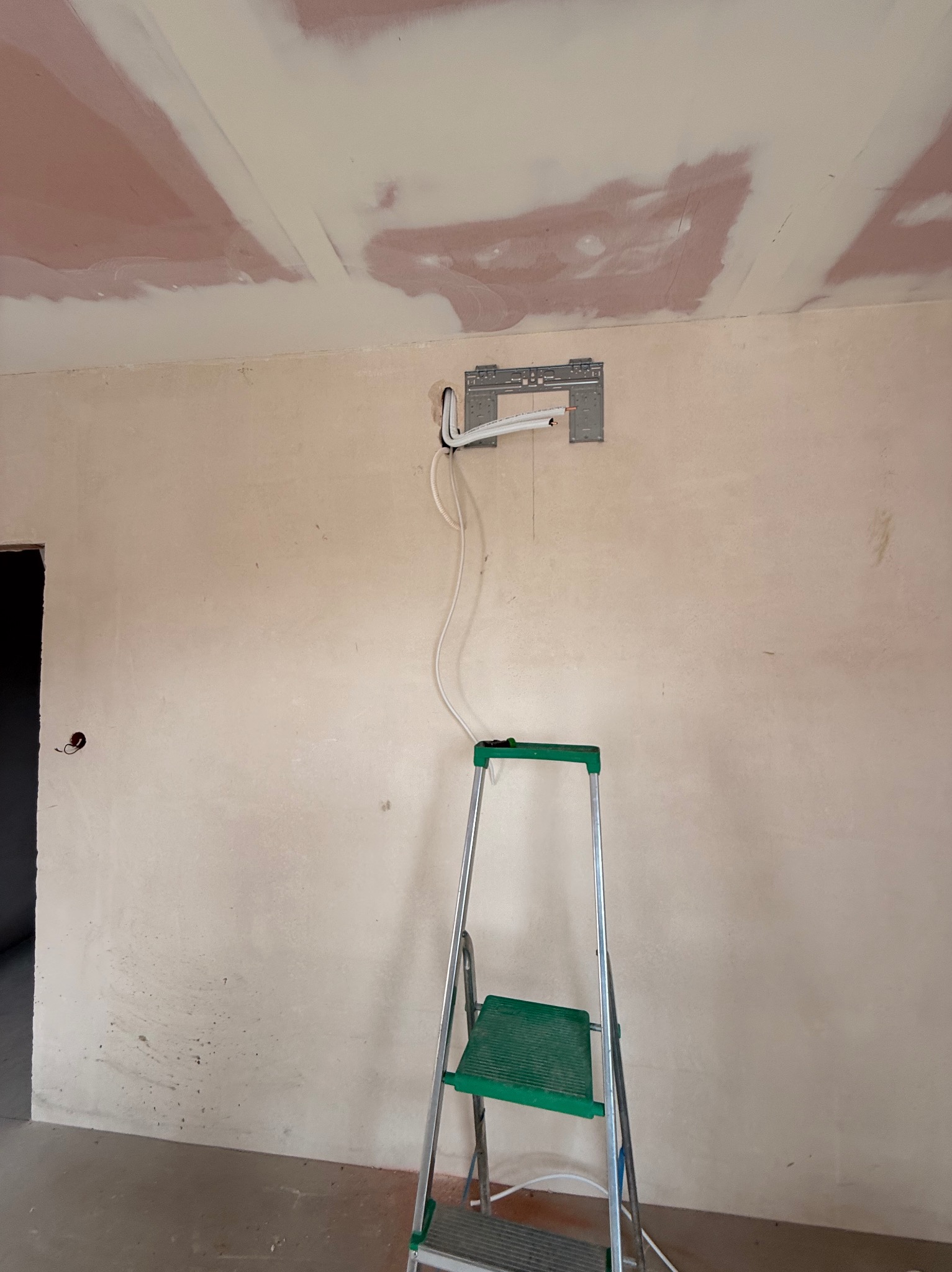 Wnętrze w trakcie montażu klimatyzacji: wspornik na ścianie, rurki, drabina. Ściana w surowym stanie, widoczne ślady po szpachlowaniu na suficie.