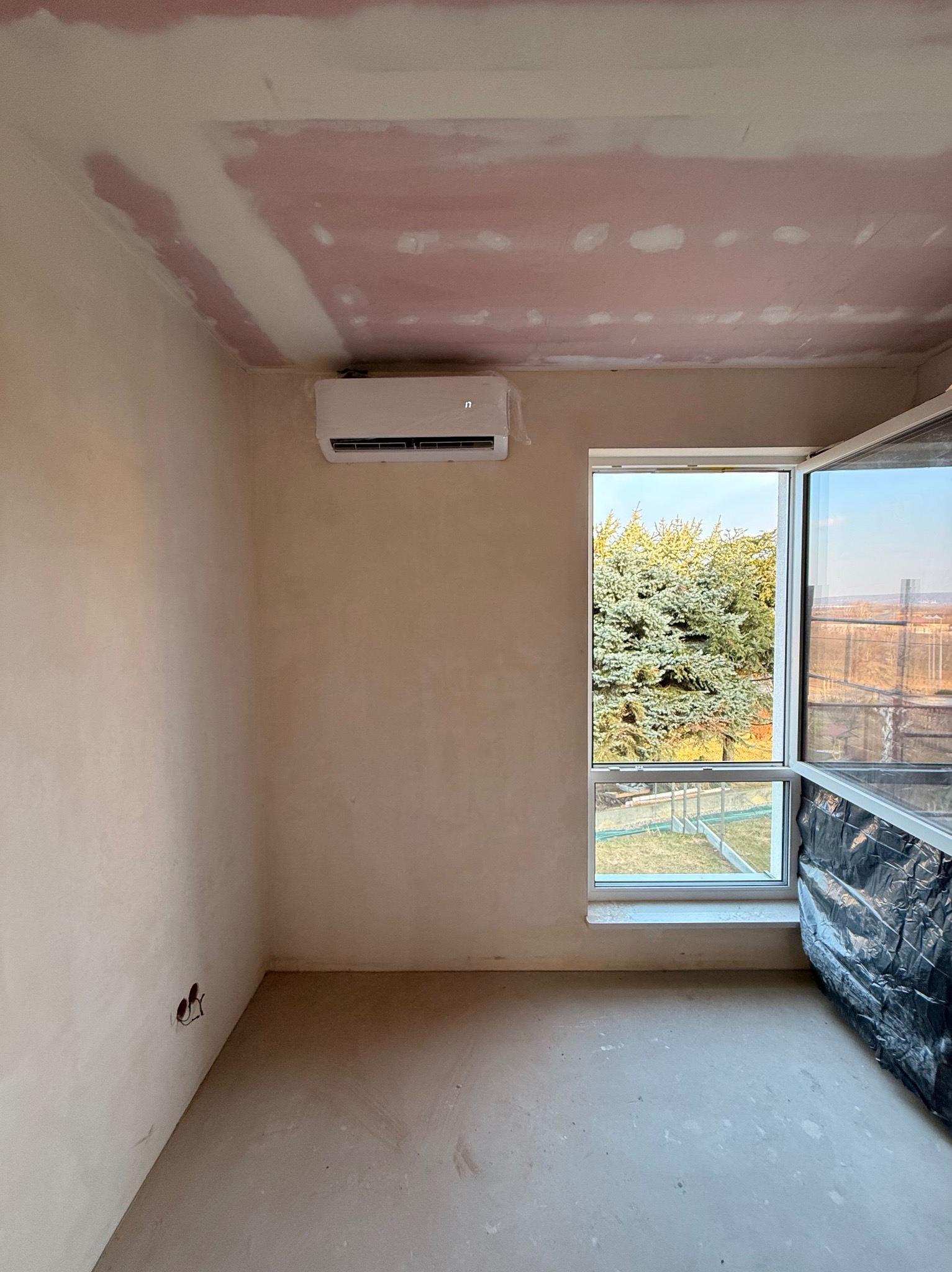 Biały klimatyzator ścienny zamontowany w surowym wnętrzu z oknem, widoczne przygotowania do wykończenia ścian i podłogi. Otwór okienny z widokiem na zieleń.