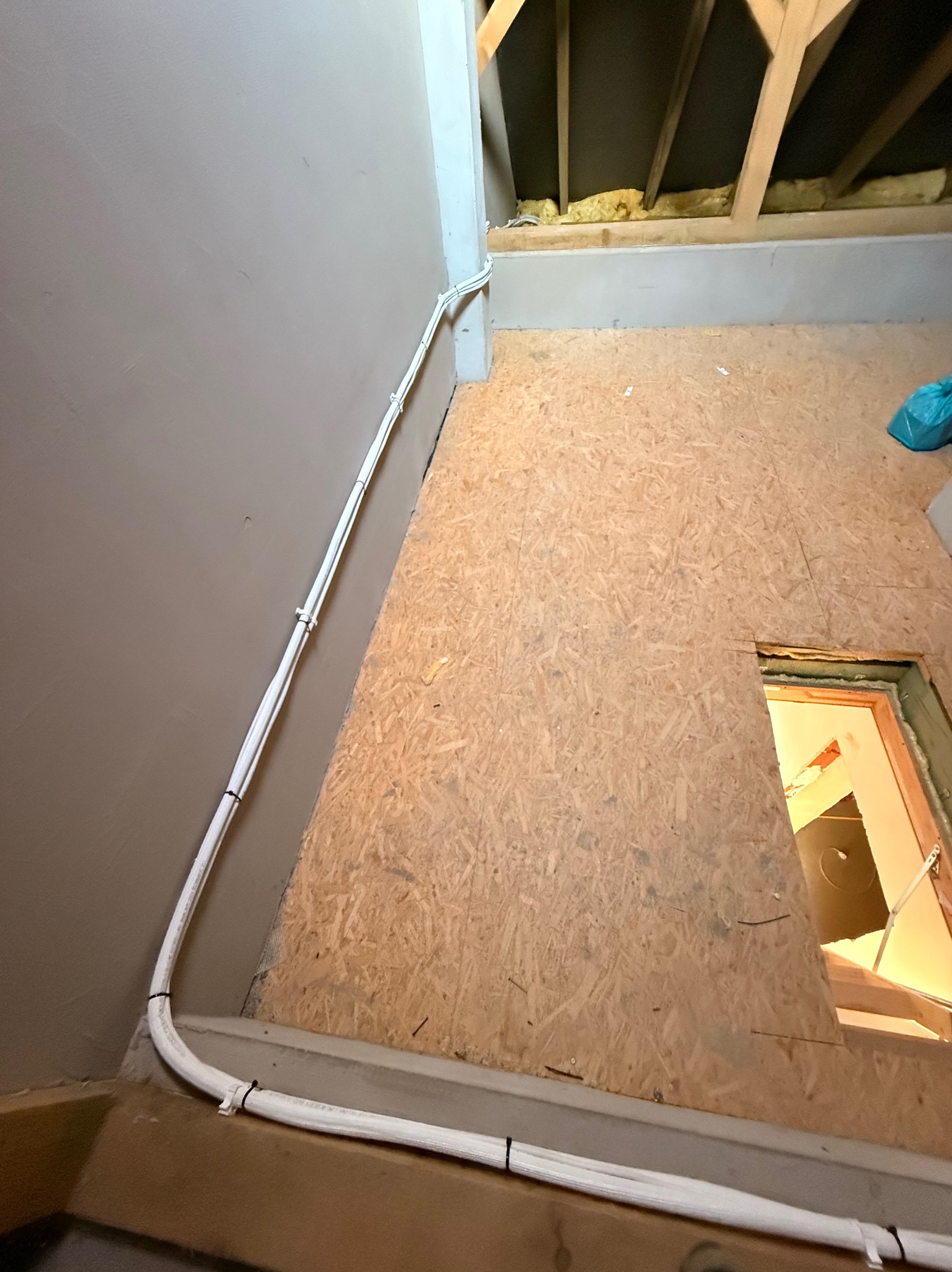 Białe kable elektryczne prowadzone wzdłuż szarej ściany i drewnianej podłogi na poddaszu, zabezpieczone czarnymi opaskami zaciskowymi. Widoczna izolacja stropu.