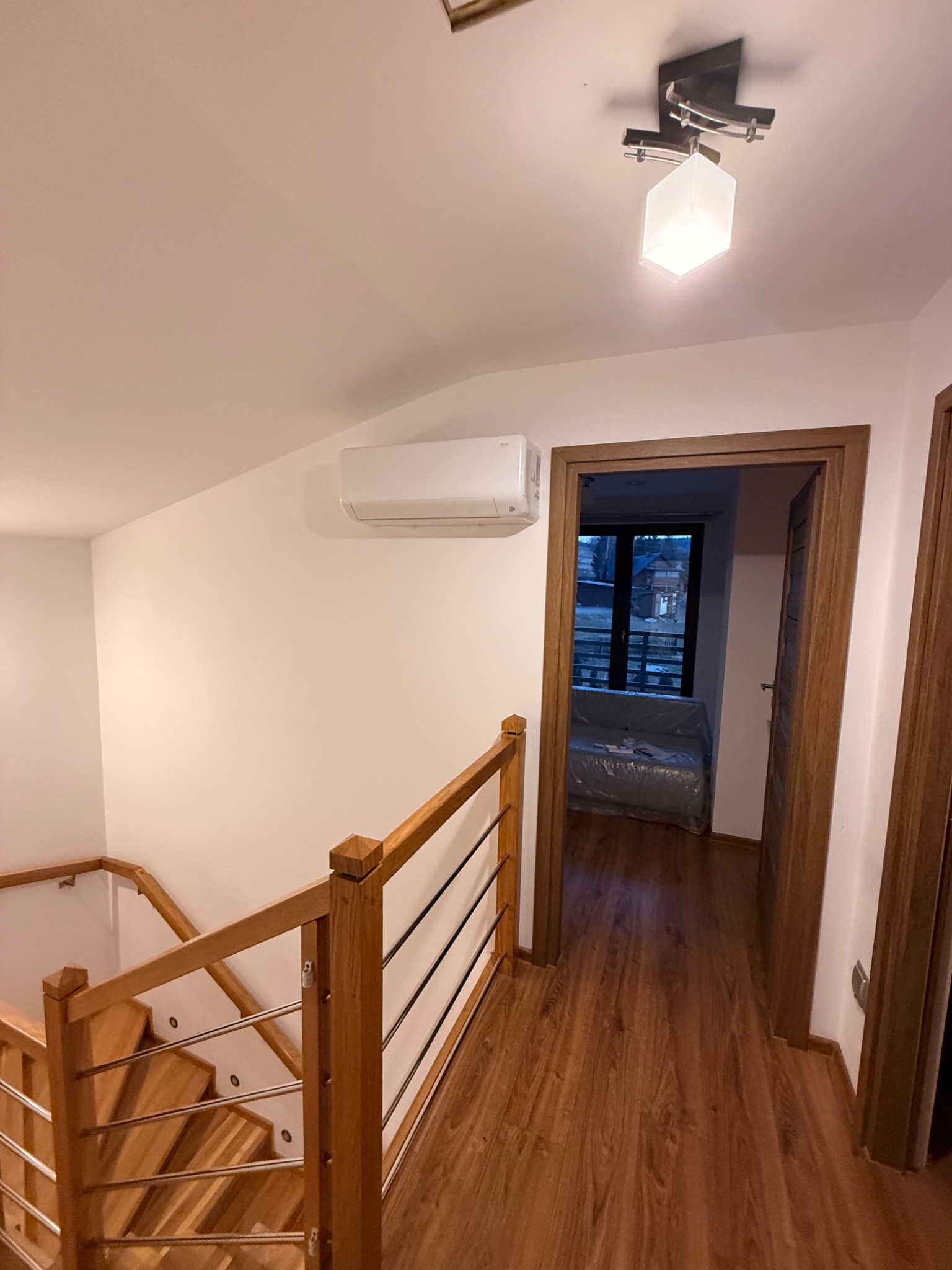 Biała klimatyzacja ścienna w jasnym wnętrzu domu, widoczna z perspektywy schodów. Drewniane wykończenia, lampa sufitowa i widok na pokój przez otwarte drzwi.