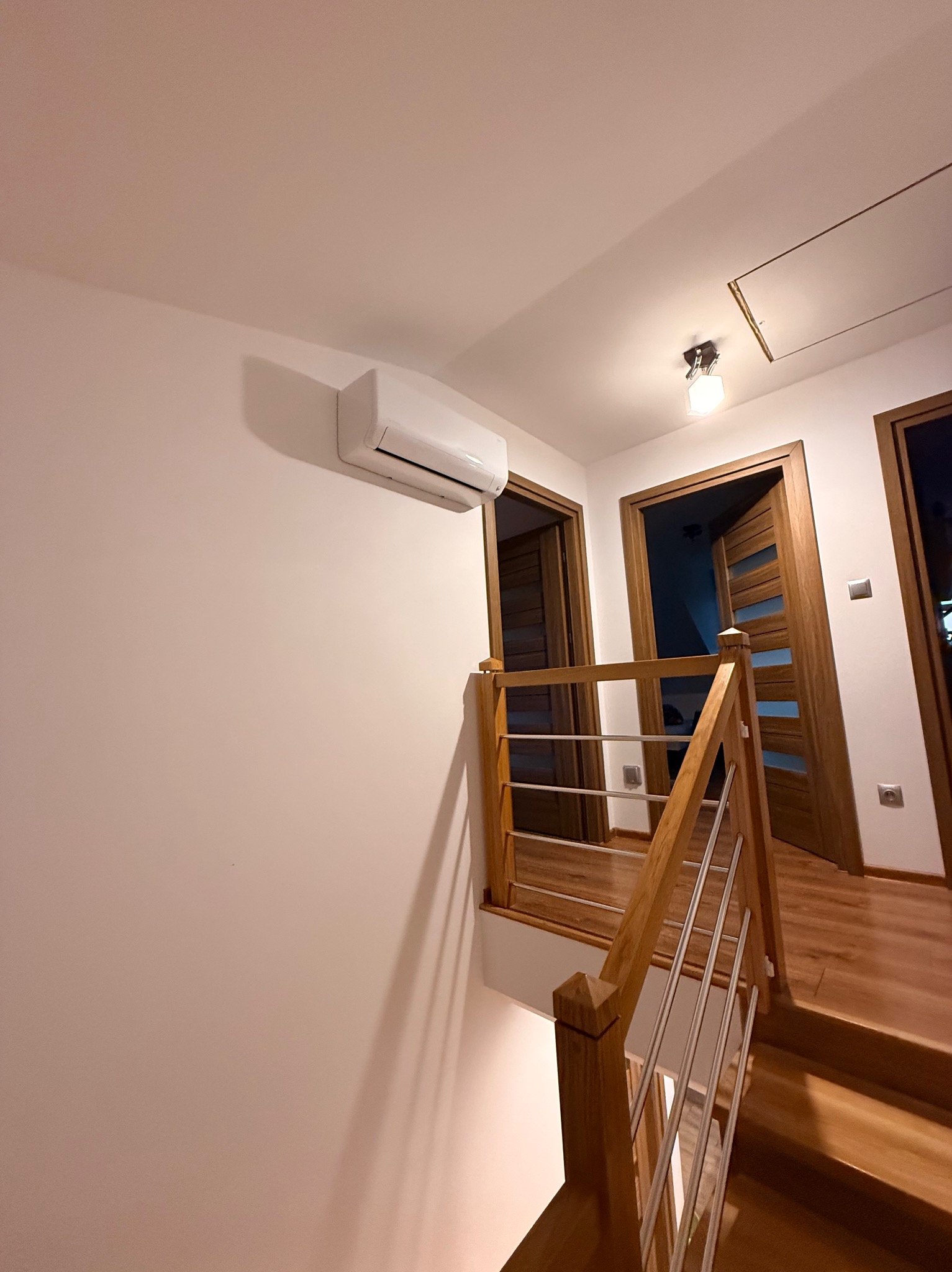 Biały klimatyzator ścienny na klatce schodowej z drewnianą balustradą i schodami. Widoczne trzy drewniane drzwi z przeszkleniami. Ciepłe oświetlenie.