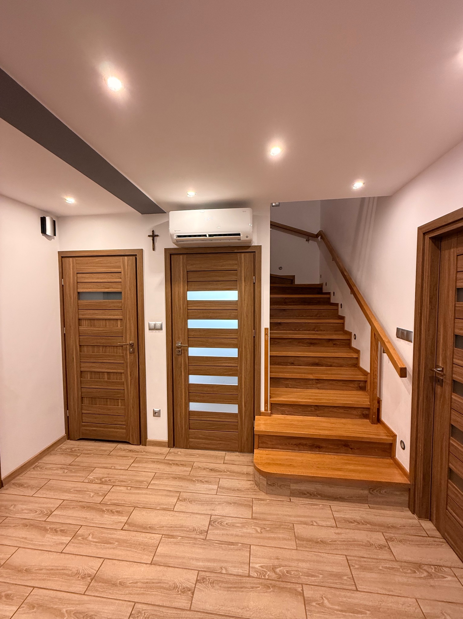 Jasne wnętrze domu z drewnianymi schodami, drzwiami i klimatyzatorem na ścianie. Widoczne oświetlenie sufitowe i dekoracyjny krzyż nad drzwiami.