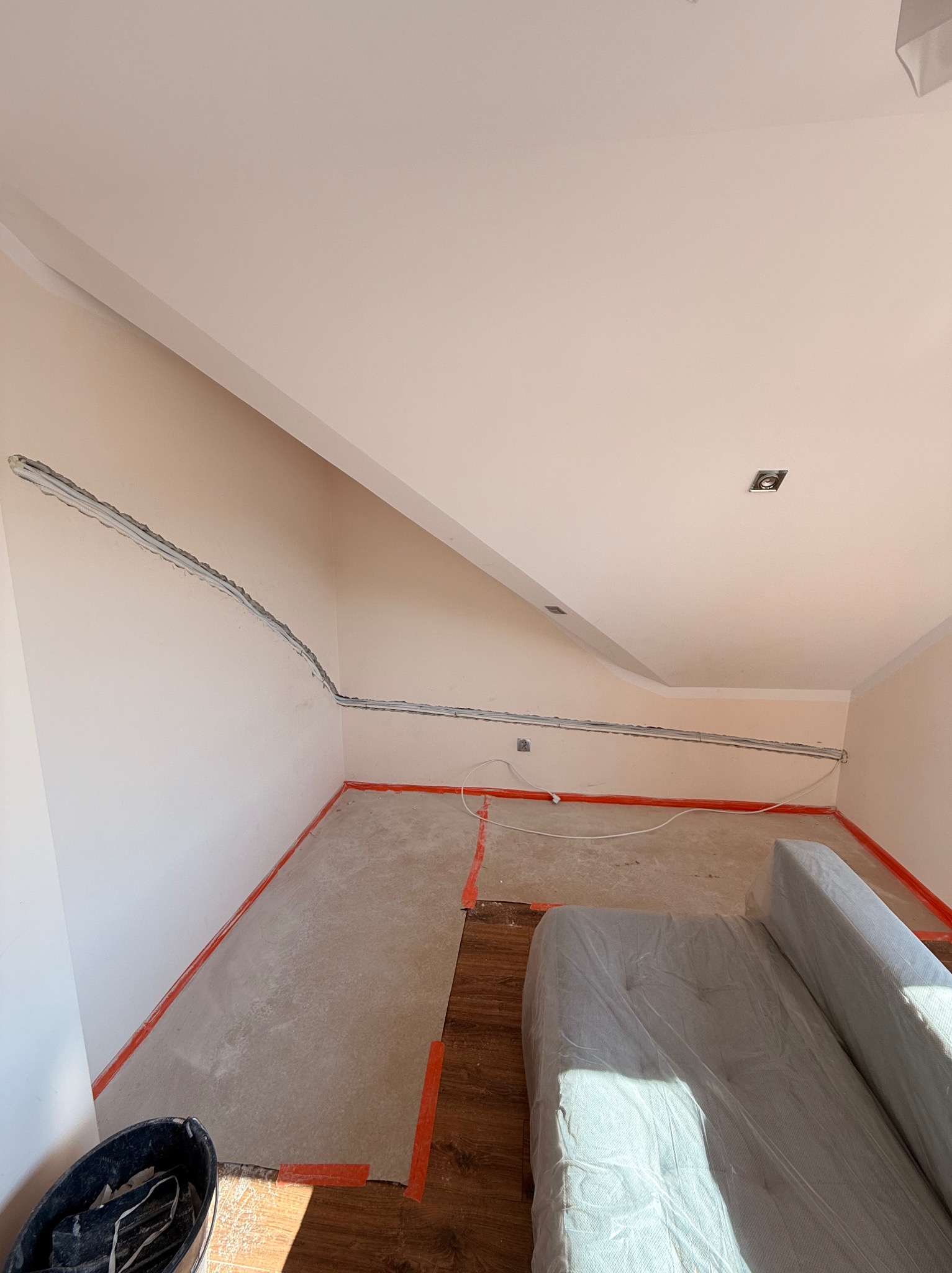 Poddasze w trakcie prac: widoczne kable w listwach na ścianach, przygotowane podłoże pod podłogę i zabezpieczone meble. Oświetlenie sufitowe w trakcie instalacji.