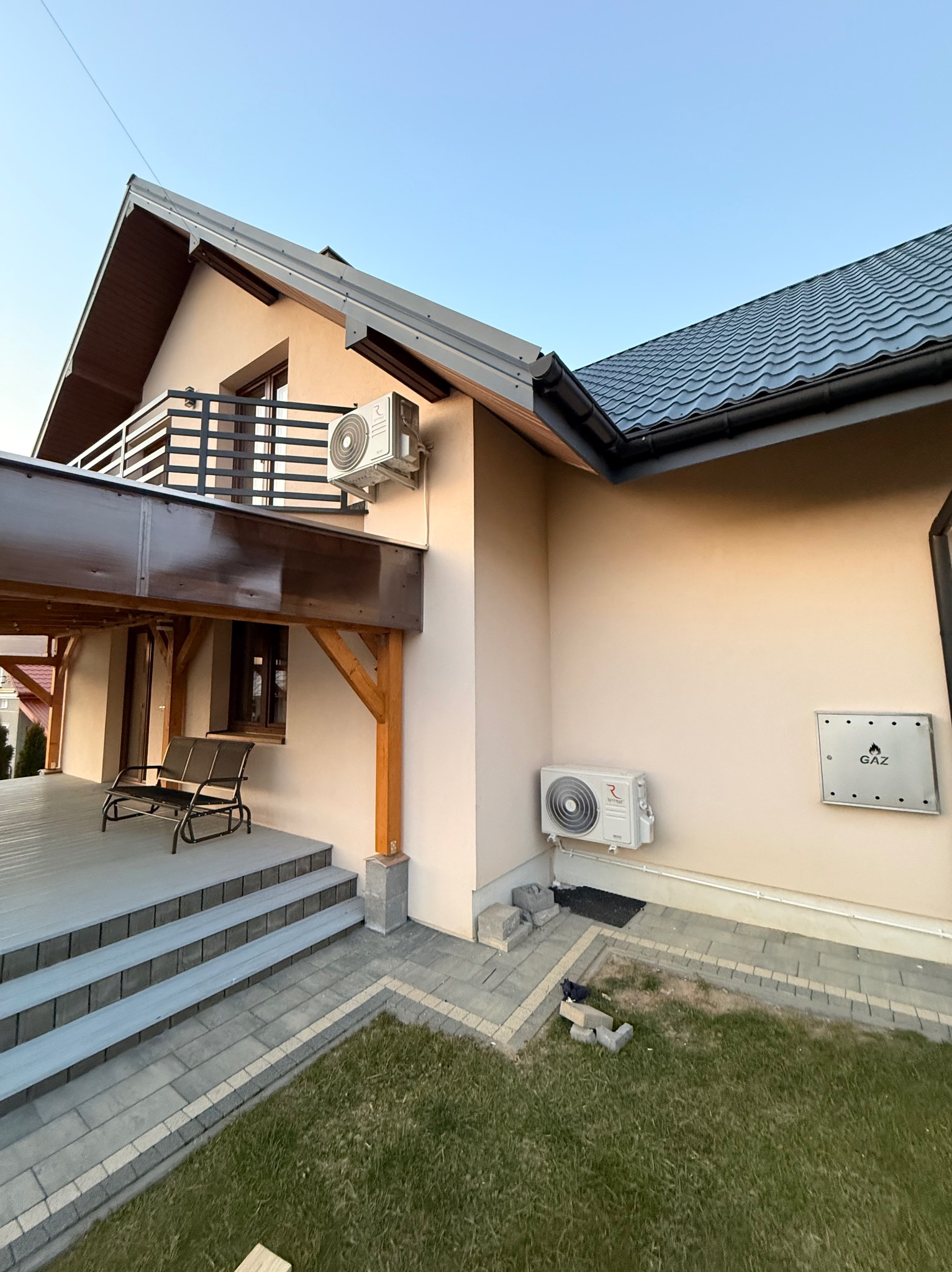 Elewacja domu z dwoma zamontowanymi klimatyzatorami i skrzynką gazową. Widoczny balkon z metalową balustradą i zadaszony taras z meblami ogrodowymi.