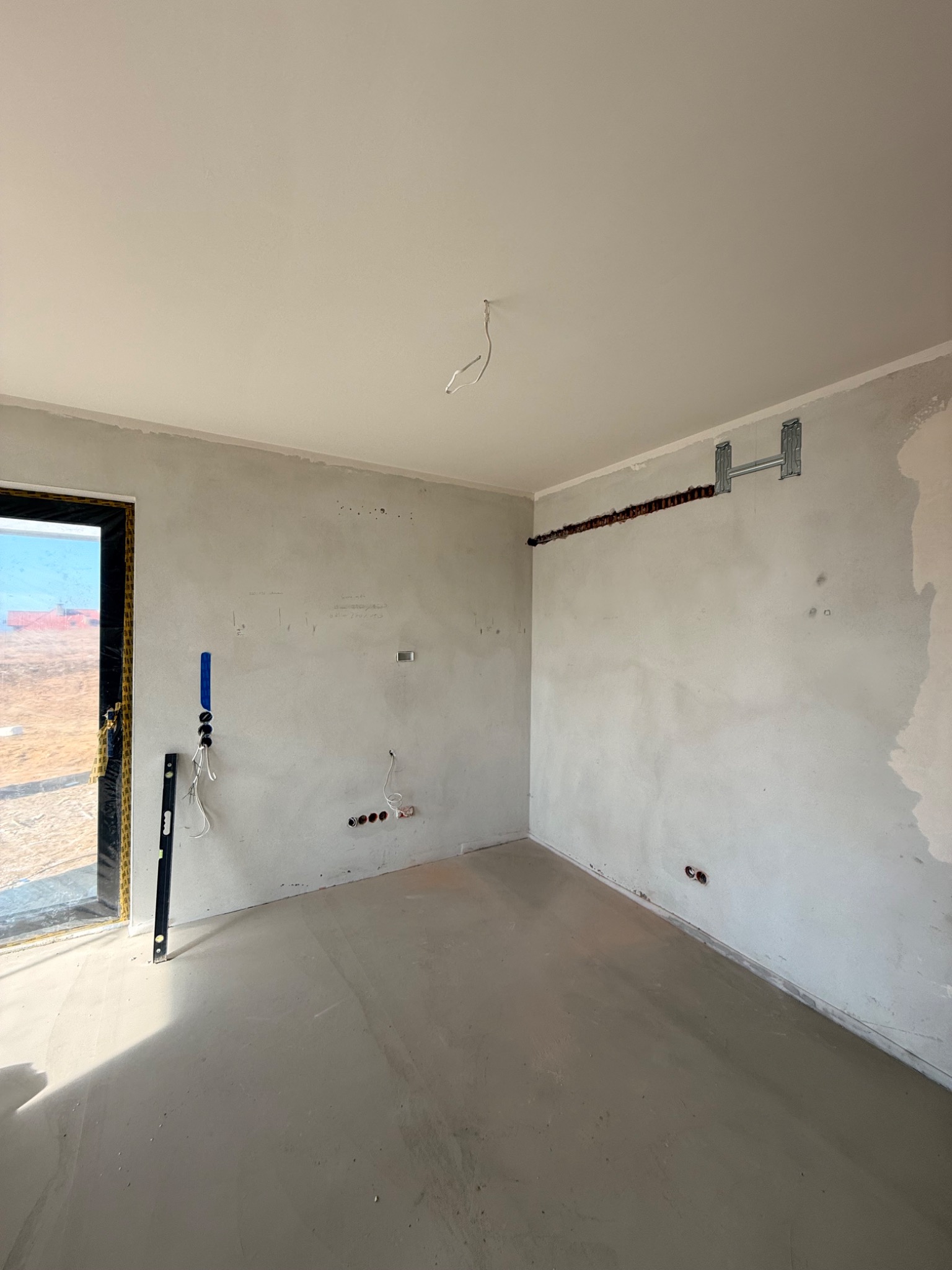 Surowy stan wnętrza: wylewka, ściany po tynku, przygotowane punkty elektryczne, uchwyt na ścianie. Widoczny poziomica przy drzwiach.