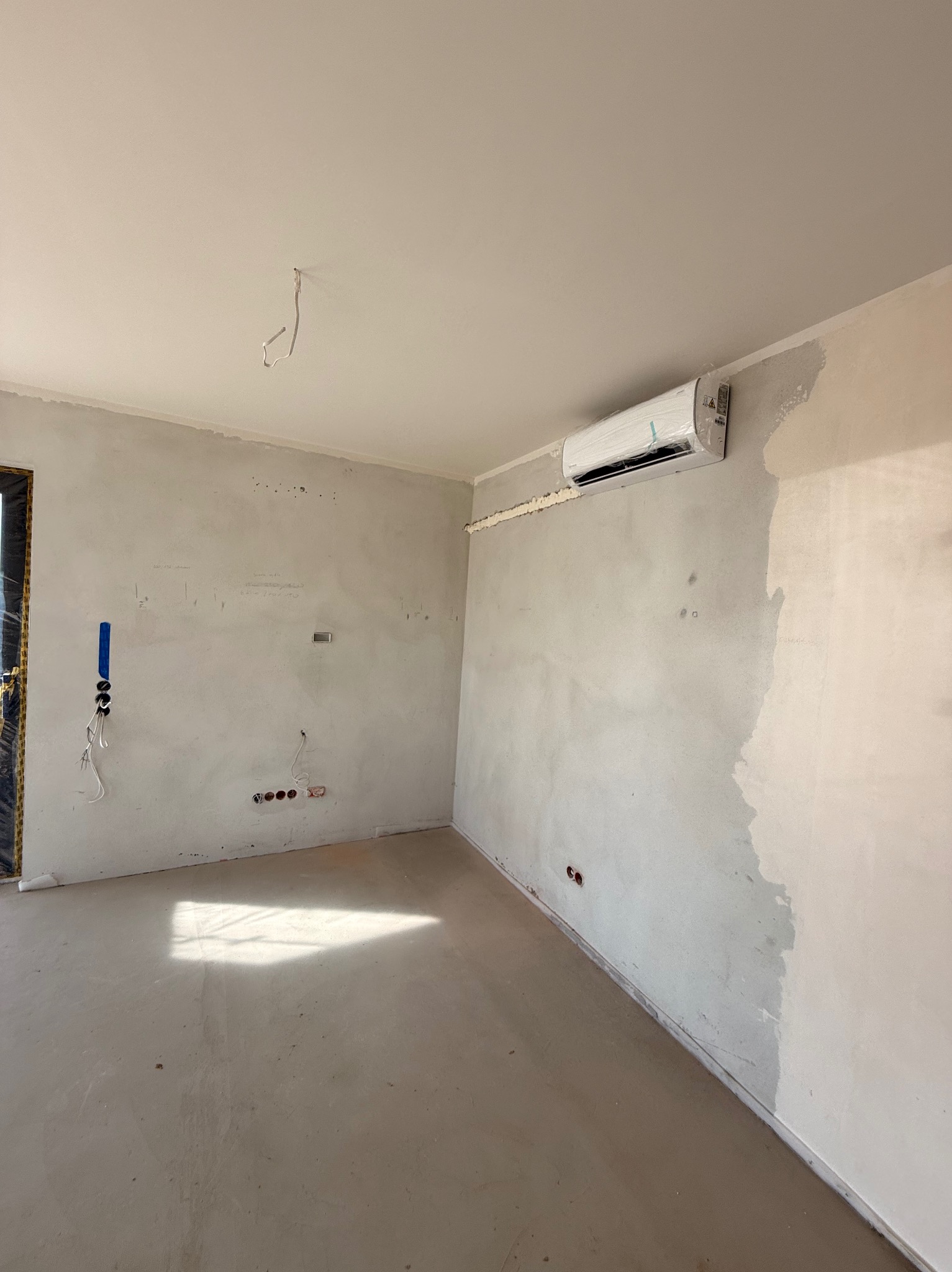 Biały klimatyzator na ścianie w surowym wnętrzu. Widoczne instalacje elektryczne i ślady po pracach wykończeniowych. Świeżo zamontowane urządzenie.