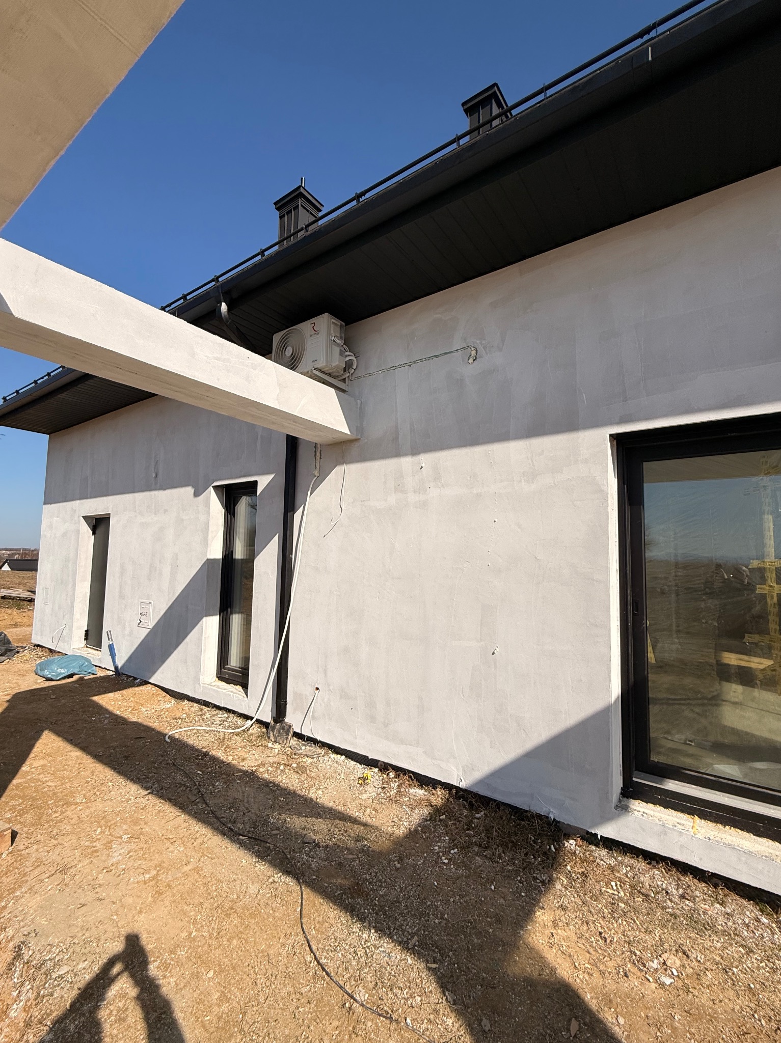 Boczny widok domu w trakcie budowy z zamontowaną jednostką zewnętrzną klimatyzacji na ścianie. Widoczne okna z czarnymi ramami i elementy konstrukcyjne.