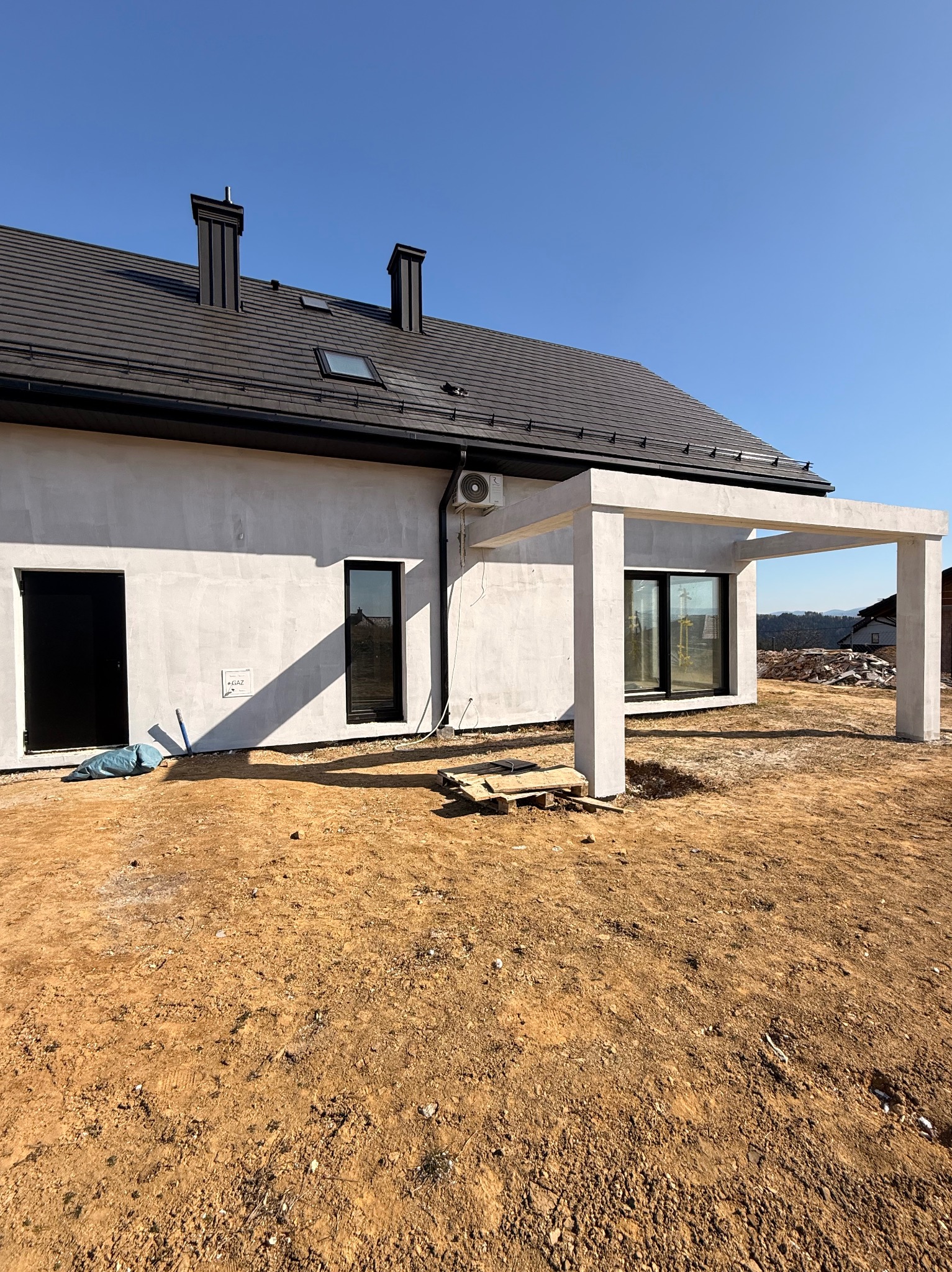 Nowoczesny dom jednorodzinny z klimatyzatorem na elewacji i betonową pergolą. Widoczne czarne okna i dachówka. Teren wokół domu w trakcie prac wykończeniowych.