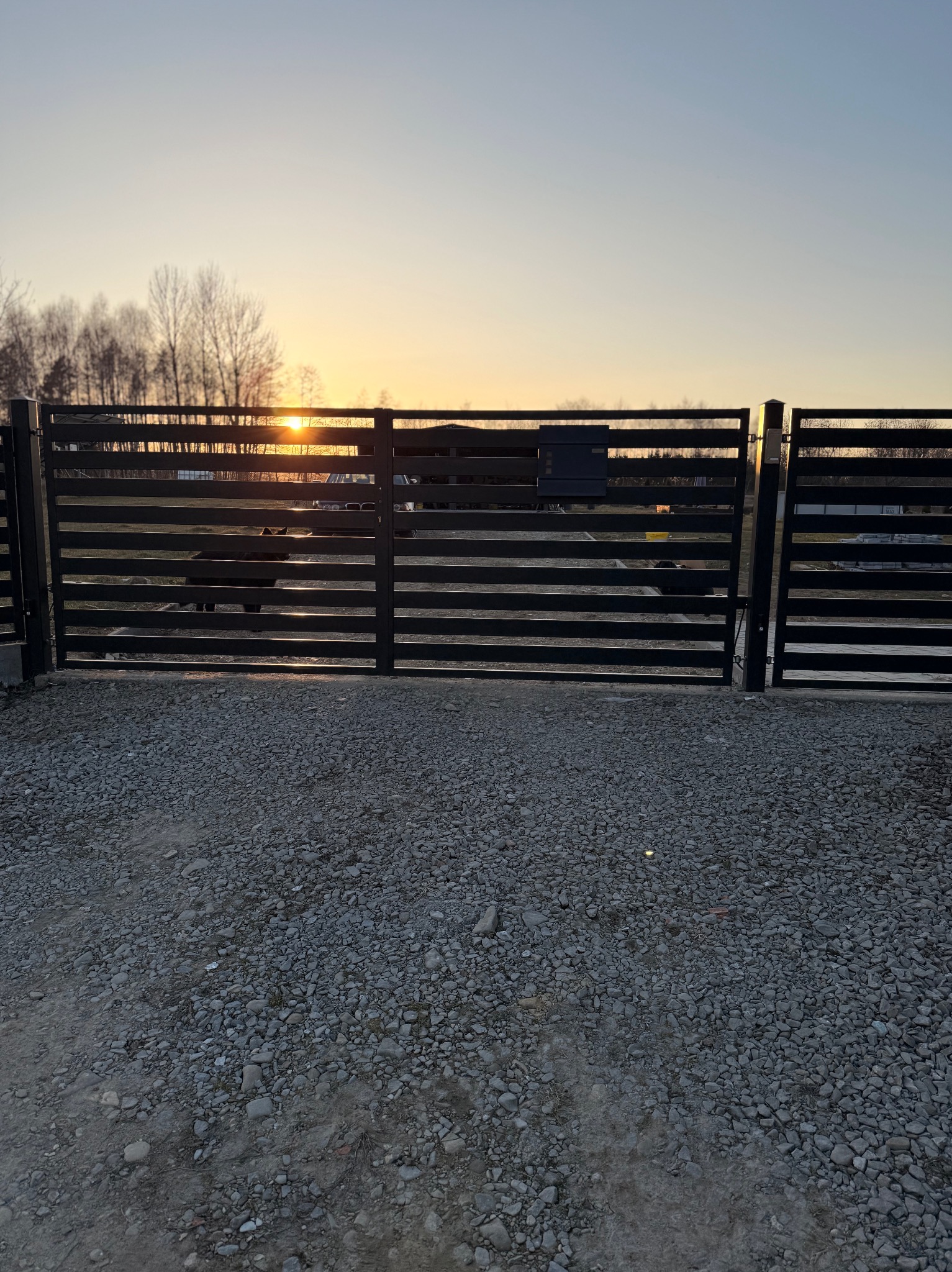 Nowoczesna, pozioma, czarna brama wjazdowa z widoczną skrzynką na listy, na tle zachodzącego słońca i drzew. Podłoże żwirowe.