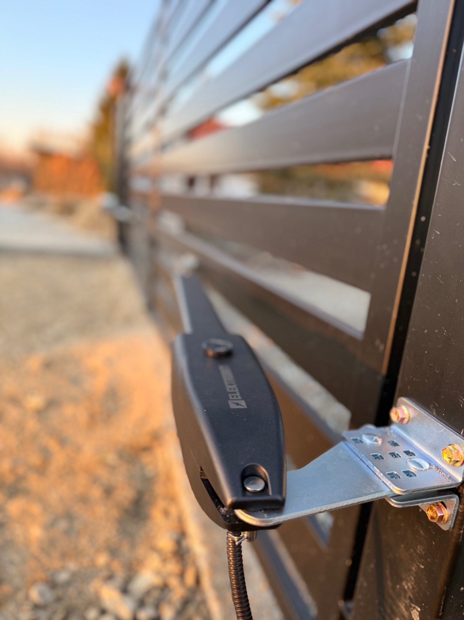 Zamknięta, nowoczesna brama posesyjna z zamontowanym napędem elektrycznym, widoczne detale mocowania i przewód. Tło rozmyte, oświetlenie popołudniowe.