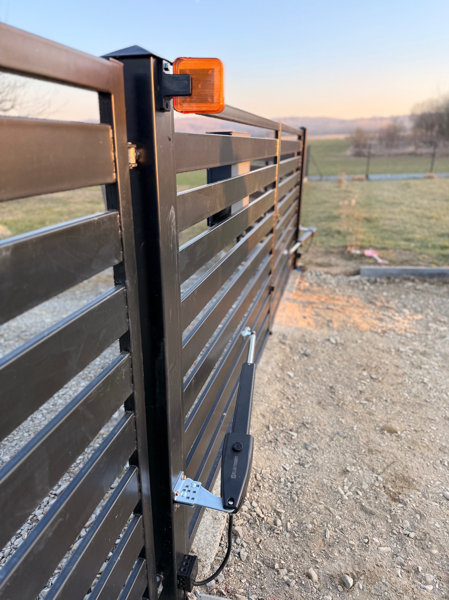 Nowoczesna, czarna brama przesuwna z automatyką i pomarańczowym światłem ostrzegawczym, zamontowana na posesji z widokiem na pagórkowaty krajobraz o zmierzchu.