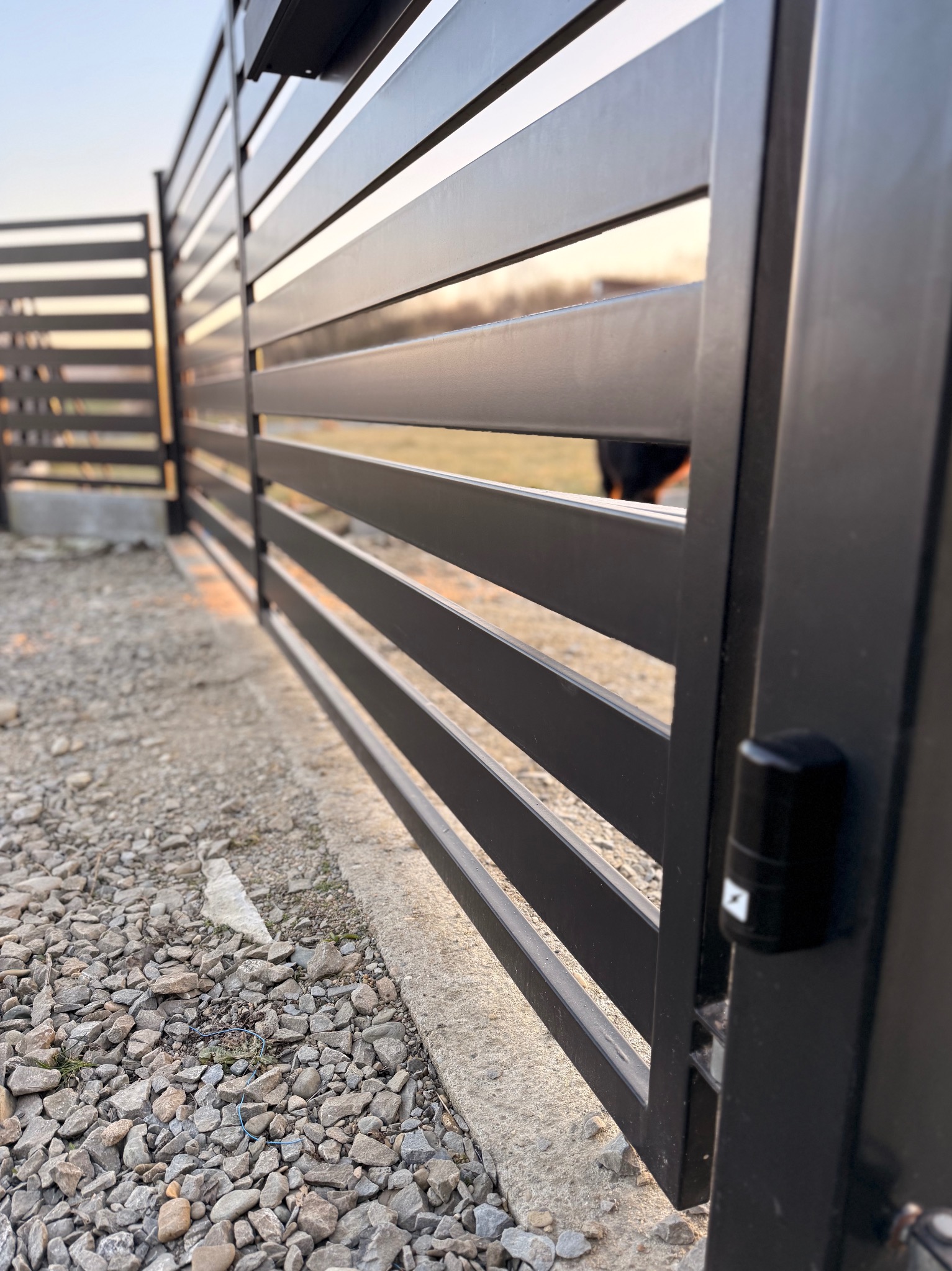 Nowoczesna, czarna brama posesyjna z poziomymi lamelami, widok z bliska na fotokomórkę. Tło rozmyte, widoczny żwir i fragment trawnika.