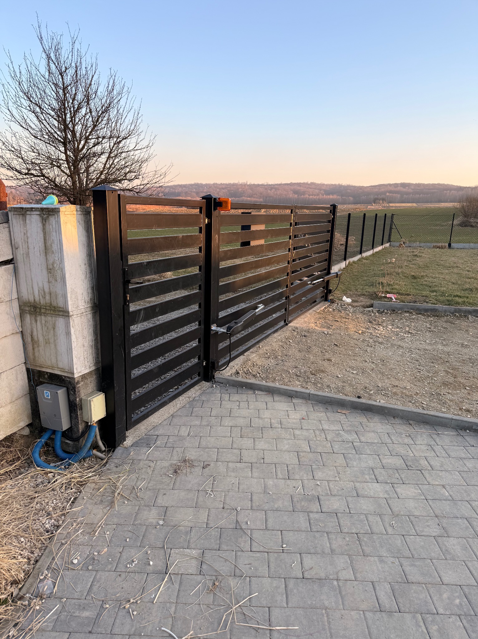 Nowoczesna, czarna brama wjazdowa z automatyką, widoczna z perspektywy kostki brukowej. W tle drzewo bez liści i zalesione wzgórza o zmierzchu.
