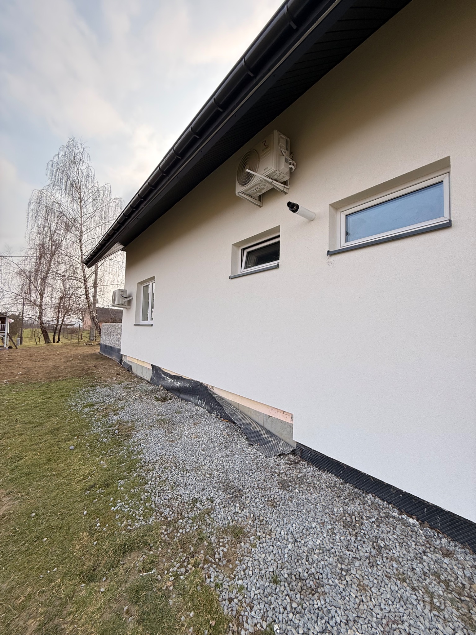 Biała ściana domu z zamontowanym klimatyzatorem i kamerą monitoringu. Widoczne okna, trawnik i drzewo bez liści na tle pochmurnego nieba.