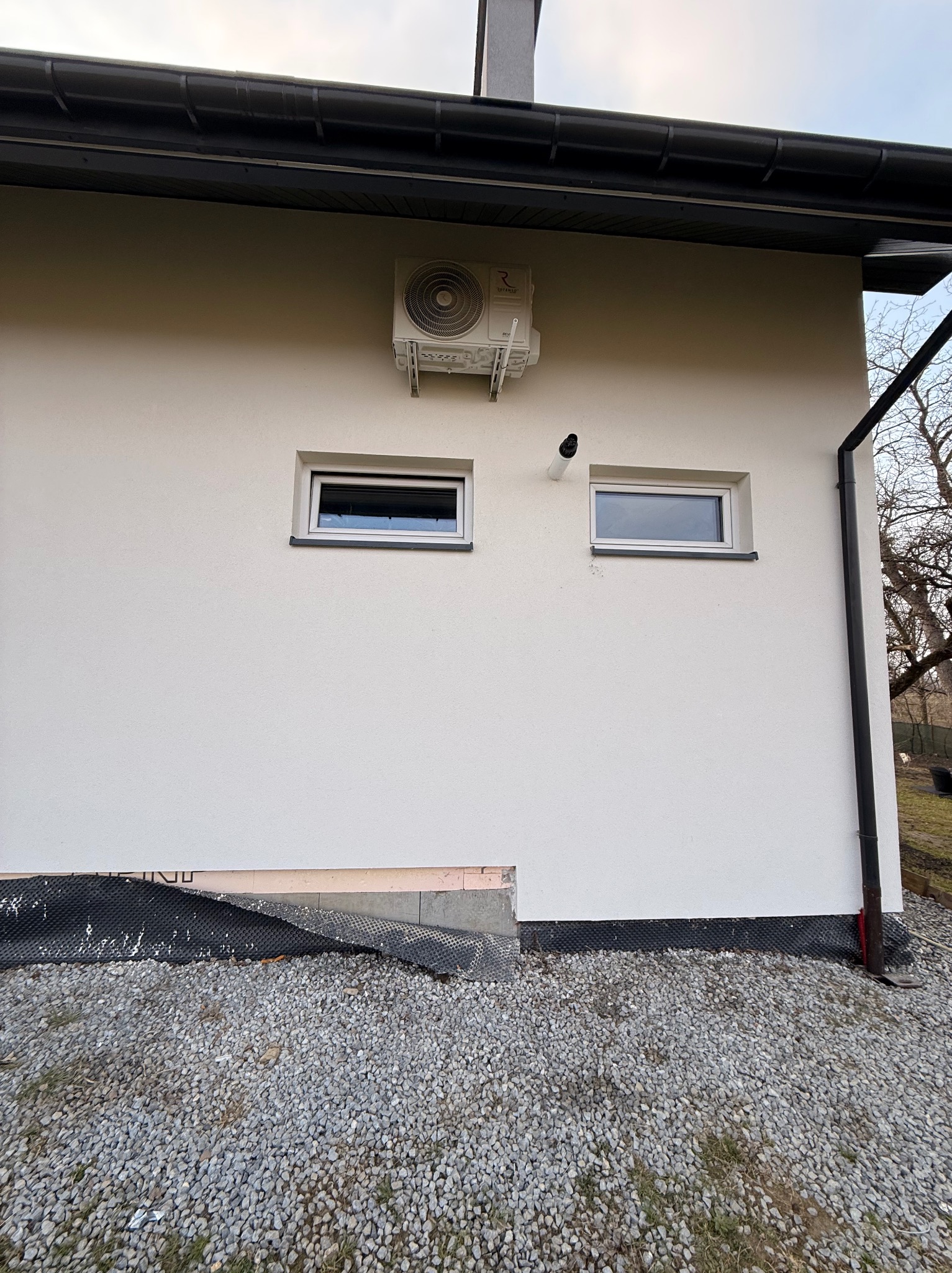 Boczny widok budynku z zamontowaną klimatyzacją na elewacji, dwa okna i fragment fundamentu z izolacją. Na dole żwir.