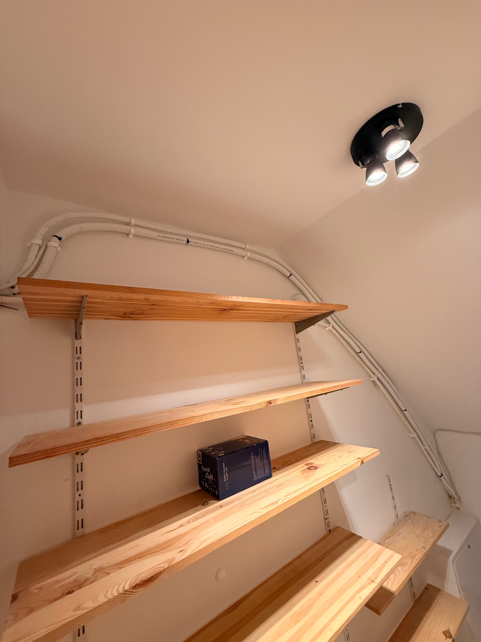 Półki z jasnego drewna zamontowane na ścianie, z widocznymi rurami instalacji elektrycznej pod sufitem i oświetleniem punktowym. Jedna półka z pudełkiem.