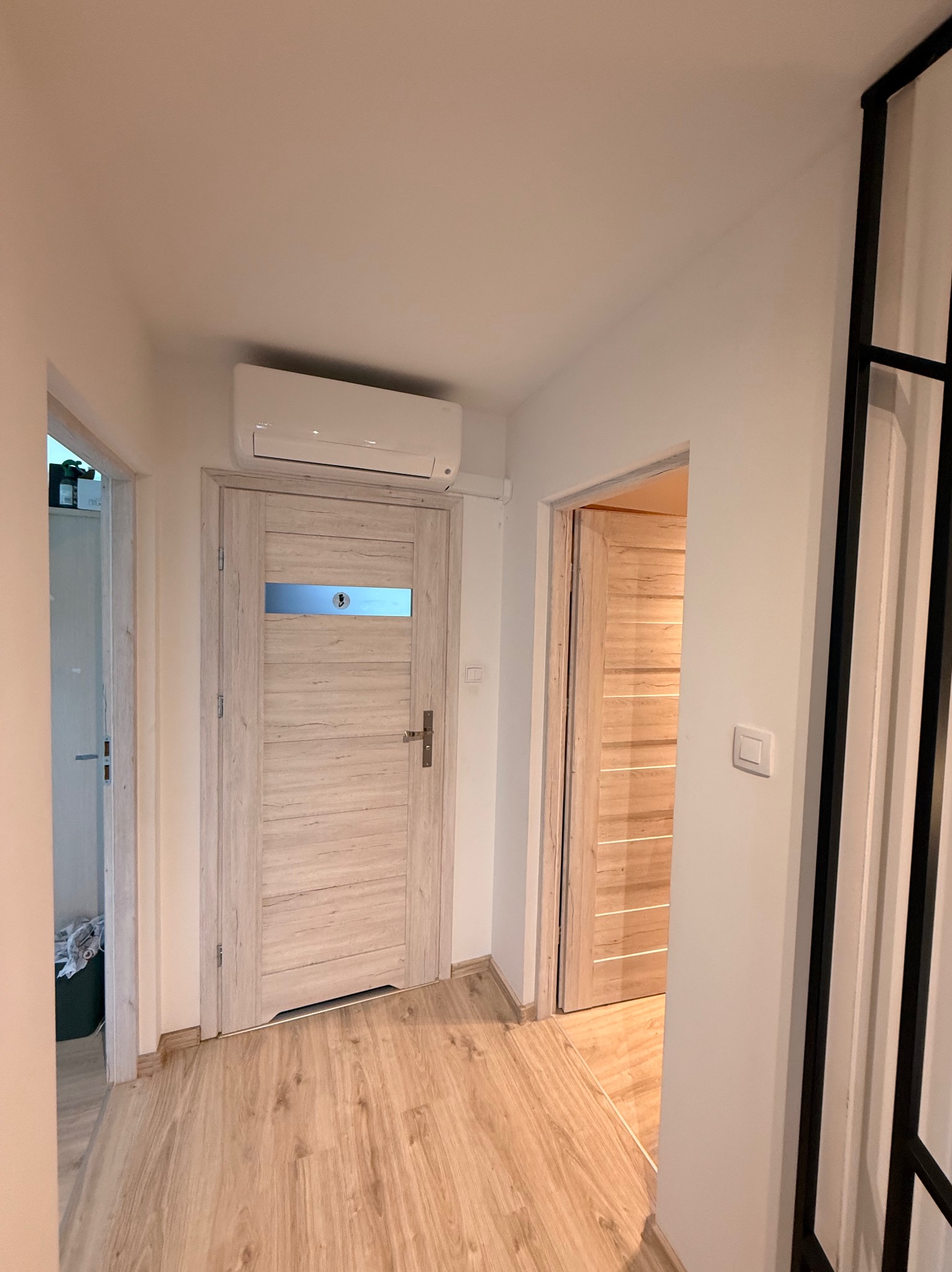 Korytarz z klimatyzatorem nad drzwiami. Widoczne dwie pary drzwi z drewnianym wykończeniem i panelami. Na ścianie włącznik światła. Podłoga wyłożona panelami.
