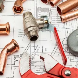 Miedziane kształtki, zawór, rury, nóż introligatorski, obcinak do rur i klucz nastawny na planie instalacji hydraulicznej.