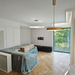 TARNA Sp.z o.o. - Jasne wnętrze po remoncie z parkietem w jodełkę, zasłoniętymi meblami, grzejnikami i nowoczesnym oświetleniem.