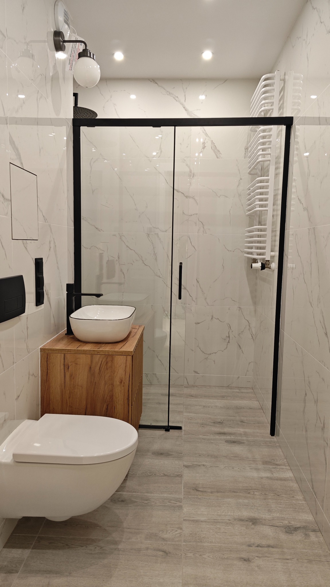 Nowoczesna łazienka z marmurowymi płytkami, kabiną prysznicową z czarną ramą, wiszącą toaletą i umywalką nablatową na drewnianej szafce.