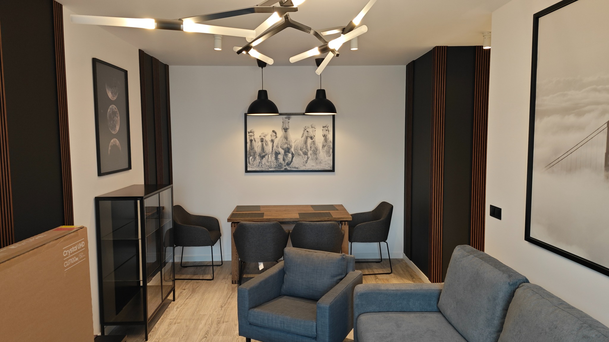 Nowoczesne wnętrze salonu z szarą sofą, drewnianym stołem i krzesłami, oświetlone designerską lampą sufitową i lampami wiszącymi, z dekoracyjnymi panelami ściennymi i obrazami w czarnych ramach.