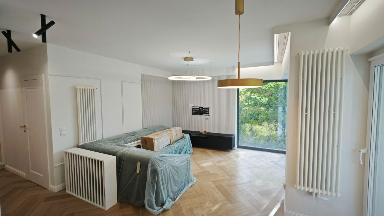 Jasne wnętrze po remoncie z parkietem w jodełkę, zasłoniętymi meblami, grzejnikami i nowoczesnym oświetleniem.