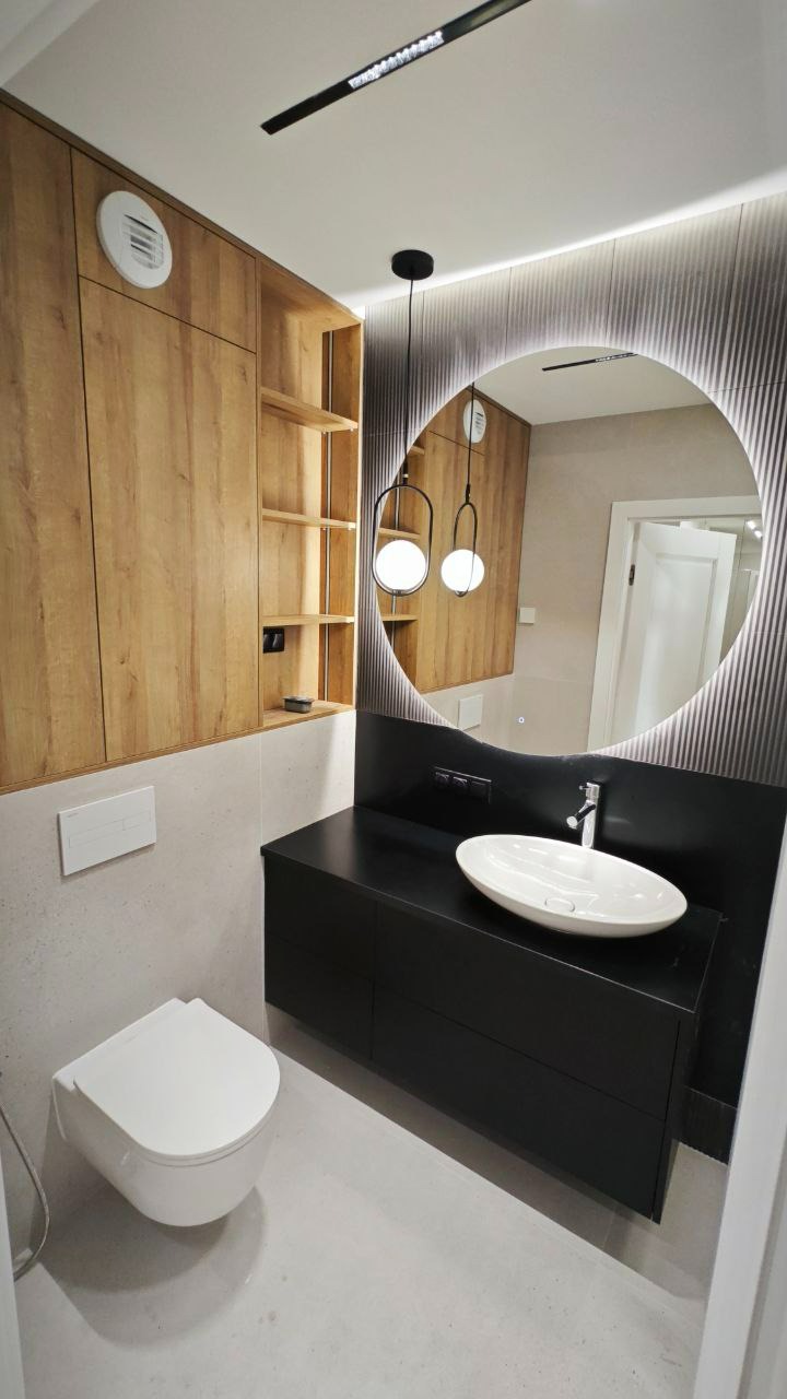 Nowoczesna łazienka z okrągłym lustrem podświetlanym LED, wiszącą szafką z czarnym blatem i owalną umywalką nablatową, wiszącą toaletą i drewnianymi szafkami z otwartymi półkami.