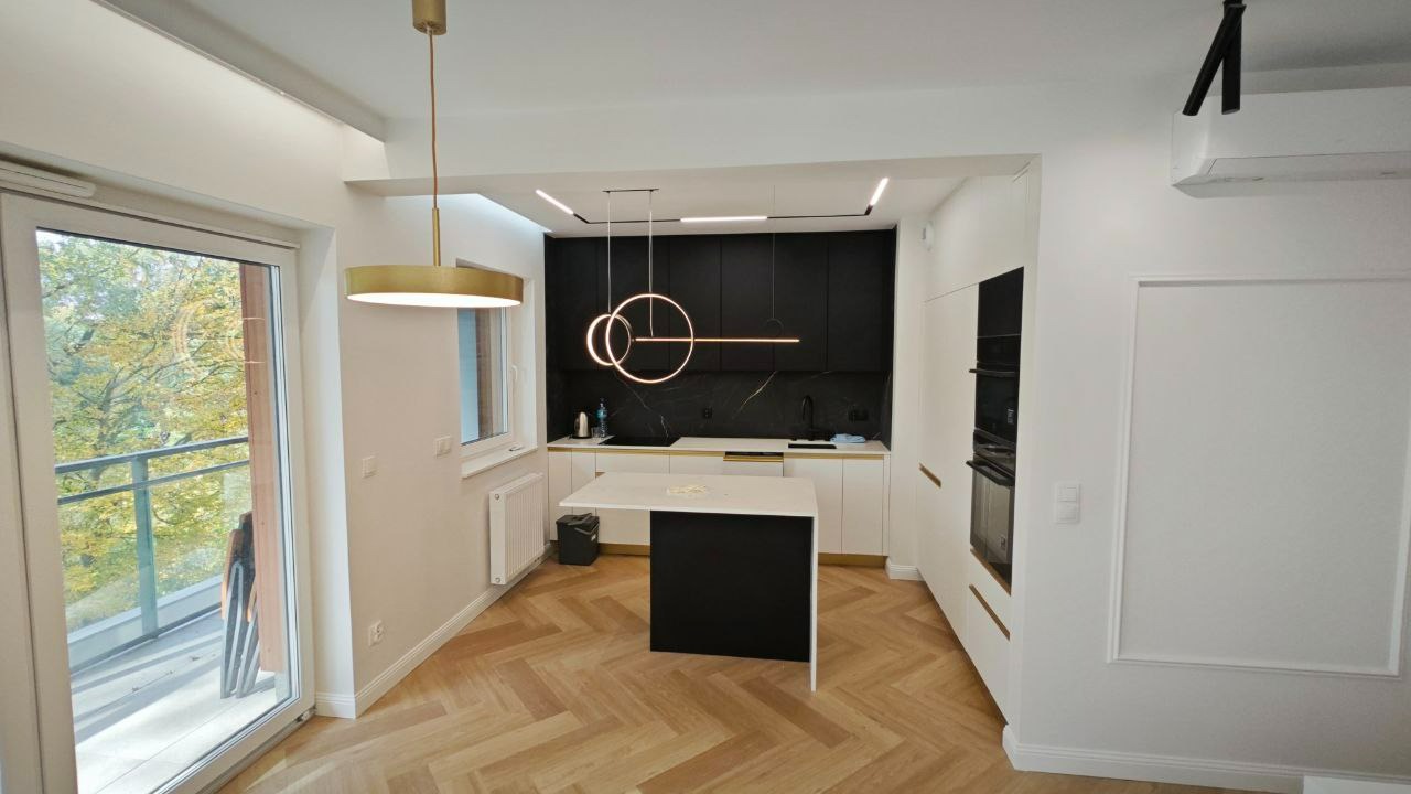 Nowoczesna kuchnia z czarnymi szafkami i marmurowym backsplashiem, oświetlona okrągłymi lampami wiszącymi, parkiet w jodełkę na podłodze, widok na balkon z zielenią za oknem.