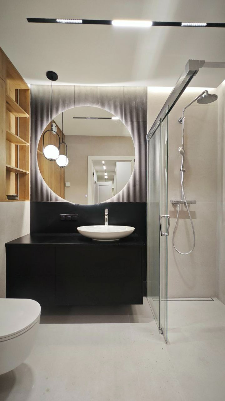 Nowoczesna łazienka z okrągłym lustrem podświetlanym LED, czarną szafką z umywalką nablatową, kabiną prysznicową ze szklanymi drzwiami i drewnianą półką.