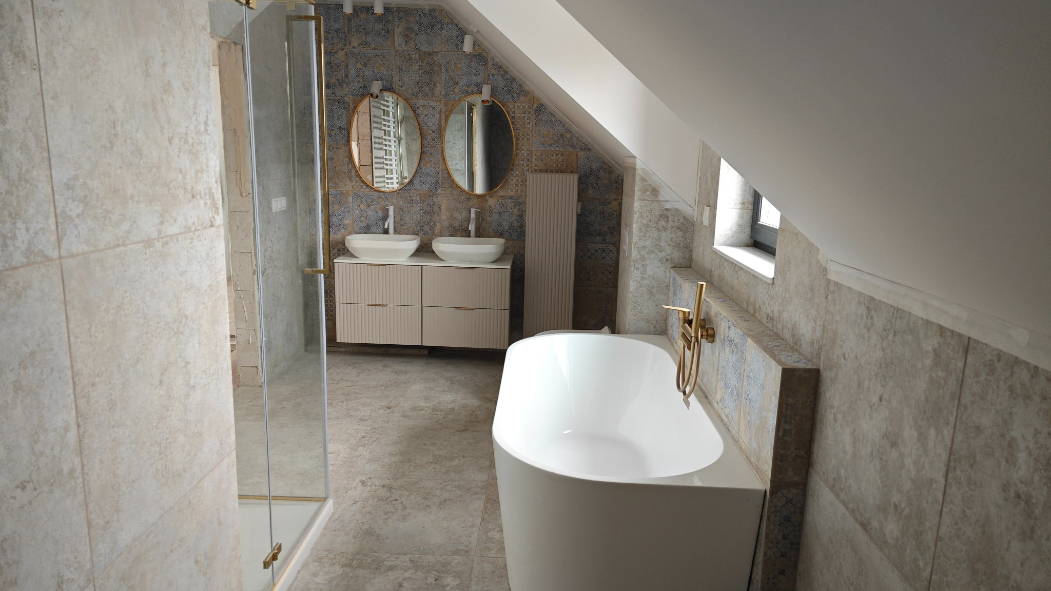 Nowoczesna łazienka na poddaszu z wolnostojącą białą wanną, beżową szafką z dwiema umywalkami, złotą armaturą i kabiną prysznicową z elementami w kolorze złota.