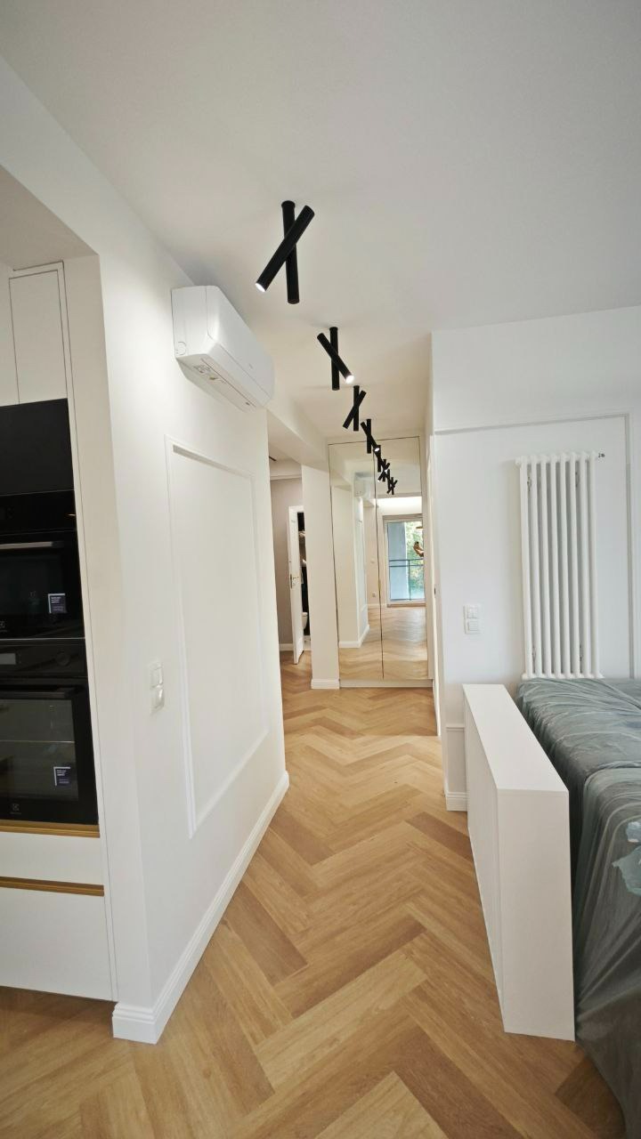 Nowoczesne wnętrze z parkietem ułożonym w jodełkę, czarnymi reflektorami sufitowymi i dużym lustrem na ścianie.