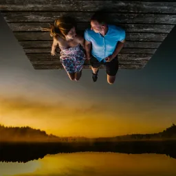 Para leżąca na drewnianym pomoście nad jeziorem o zmierzchu, odbicie drzew w wodzie tworzy surrealistyczny efekt.