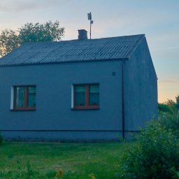 WERBA SPÓŁKA Z OGRANICZONĄ ODPOWIEDZIALNOŚCIĄ - Niewielki, szary dom z dwoma oknami i panelem słonecznym w ogrodzie, widocznym zza krzewów.