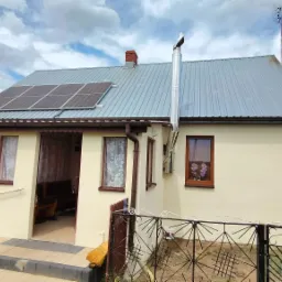 Dom jednorodzinny z panelami słonecznymi na dachu, kominem z atrapą bociana i anteną telewizyjną, ogrodzony metalowym płotem.