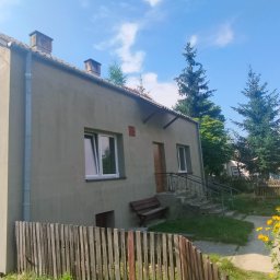 WERBA SPÓŁKA Z OGRANICZONĄ ODPOWIEDZIALNOŚCIĄ - Dwukondygnacyjny dom jednorodzinny w Warszawie z szarą elewacją, widoczny zza drewnianego płotu i kwitnących żółtych kwiatów, zarośnięty dach. Na ścianie widoczna mała czerwona tabliczka.