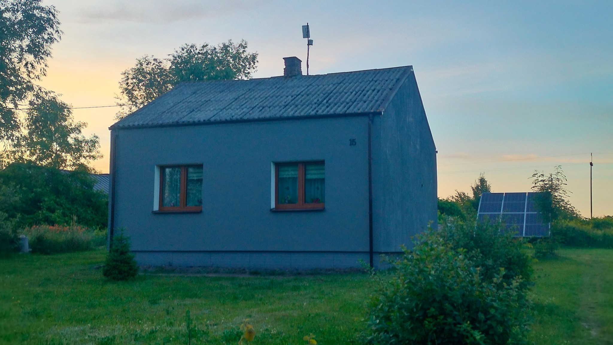 Niewielki, szary dom z dwoma oknami i panelem słonecznym w ogrodzie, widocznym zza krzewów.