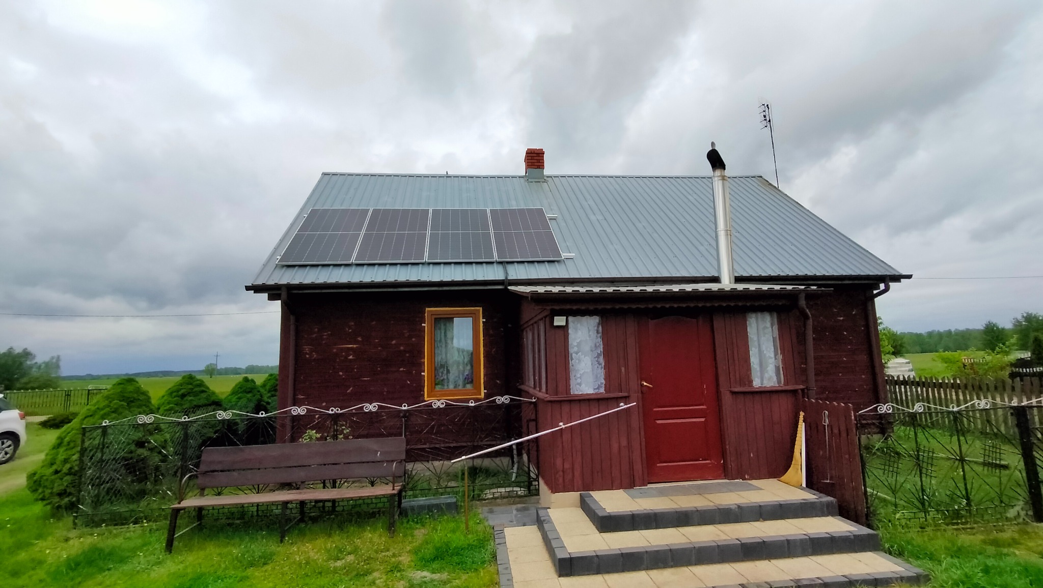 Ciemnobrązowy dom z szarą blachodachówką i panelami słonecznymi na dachu, widok z przodu, pochmurne niebo.