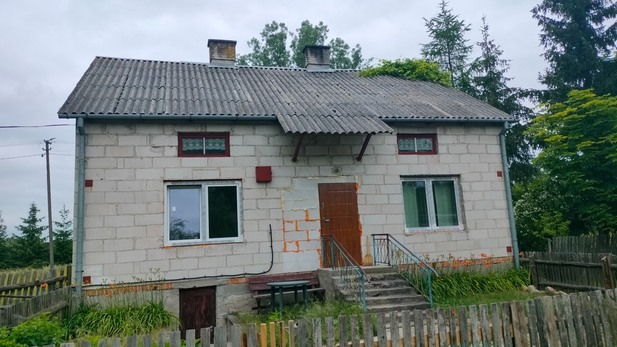 Dom jednorodzinny z szarej cegły z widocznymi śladami prac remontowych przy drzwiach wejściowych, porośnięty zielenią dach, drewniane okna i stara dachówka.