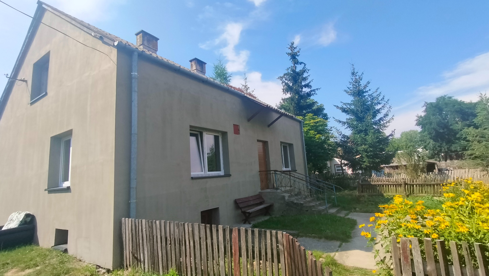 Dwukondygnacyjny dom z szarą elewacją, widoczny z boku, z drewnianym płotem i żółtymi kwiatami na pierwszym planie, oraz drzewami w tle.