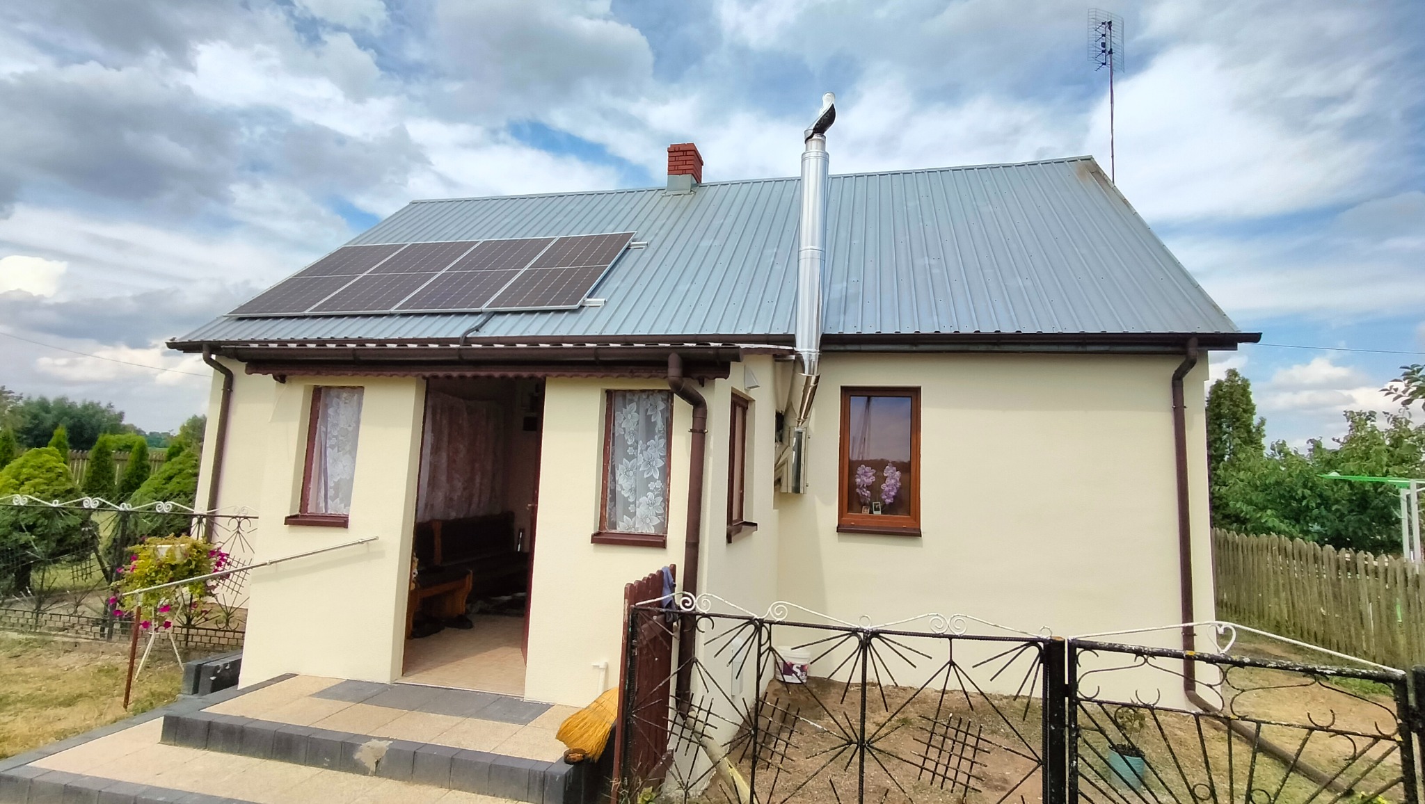 Dom jednorodzinny z panelami słonecznymi na dachu, kominem z atrapą bociana i anteną telewizyjną, ogrodzony metalowym płotem.