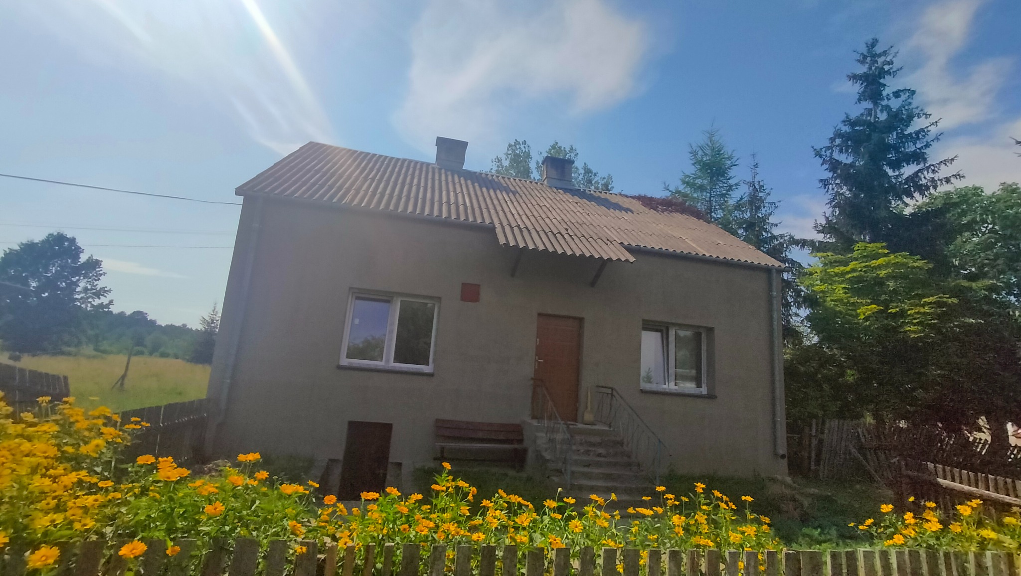 Starszy, dwukondygnacyjny dom z szarą elewacją i uszkodzonym dachem z eternitu, otoczony drewnianym płotem i kwitnącymi żółtymi kwiatami, widok w słoneczny dzień.